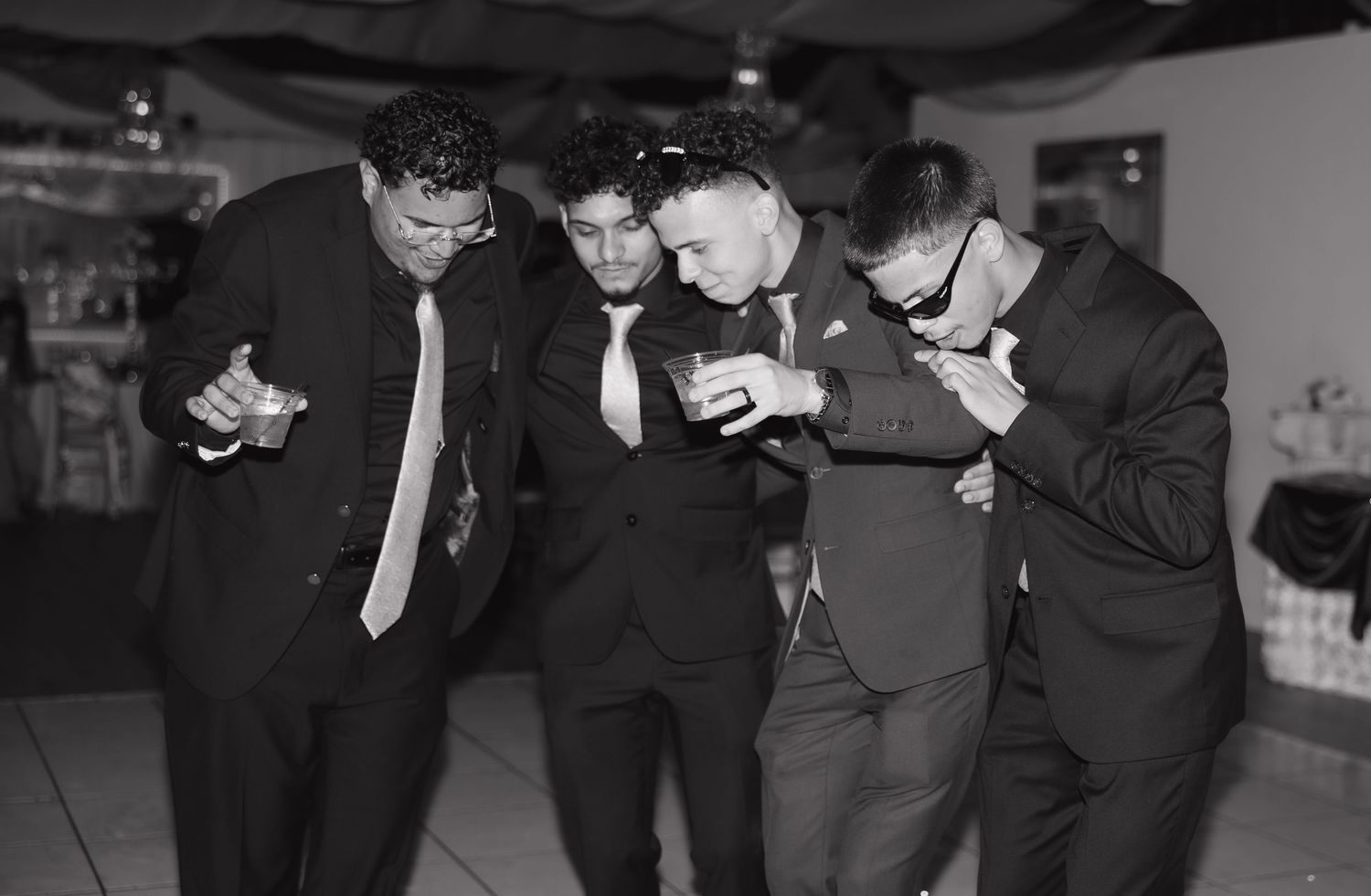 Four men in suits and ties share a joyful moment on a dance floor at a formal event in black and white.