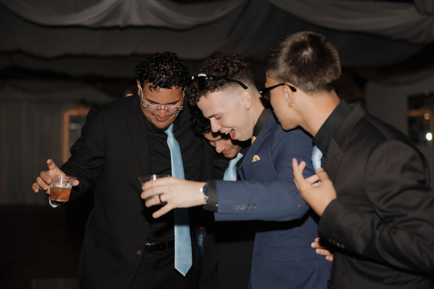 Three men in suits laughing and dancing together at a formal event, holding drinks.
