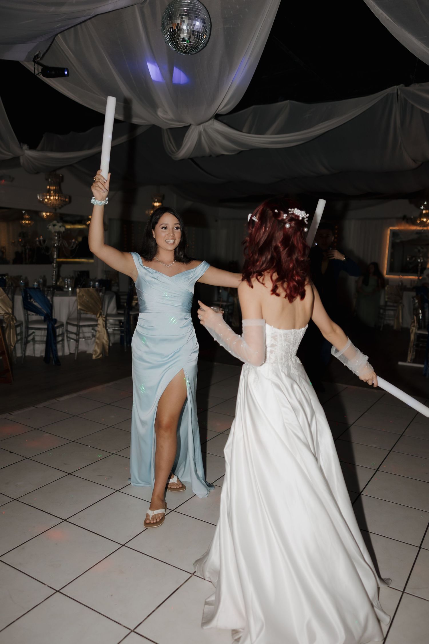 Bride in white gown and bridesmaid in blue dress dancing with light wands at elegant wedding reception.