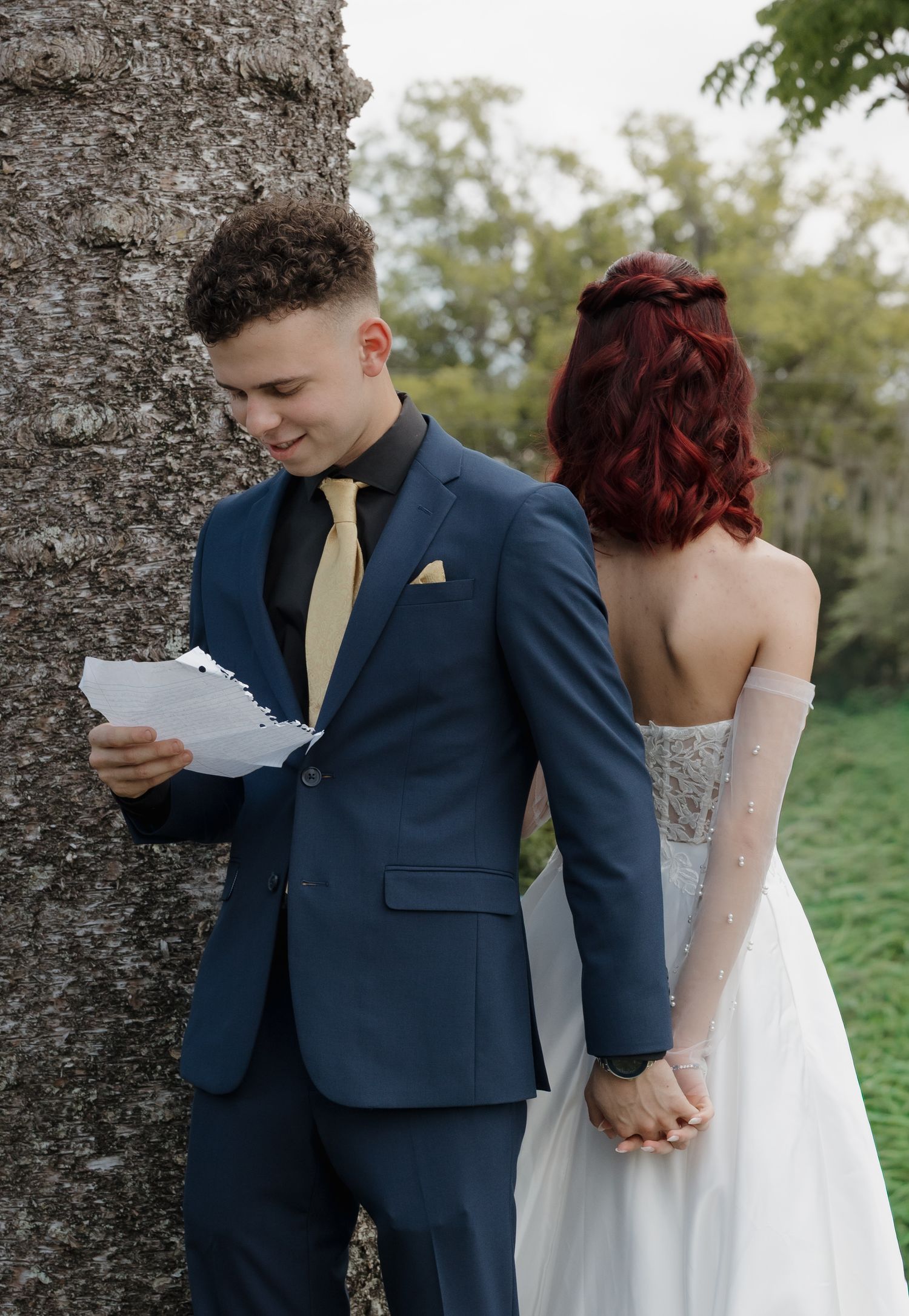 Groom in navy suit reads vows beside tree while bride in white strapless gown holds his hand outdoors.