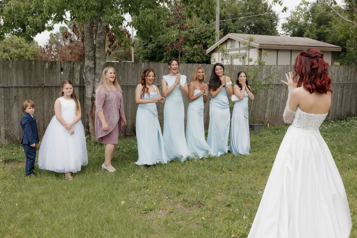 Bride in white gown faces bridesmaids in light blue dresses and flower girl on green lawn near wooden fence.