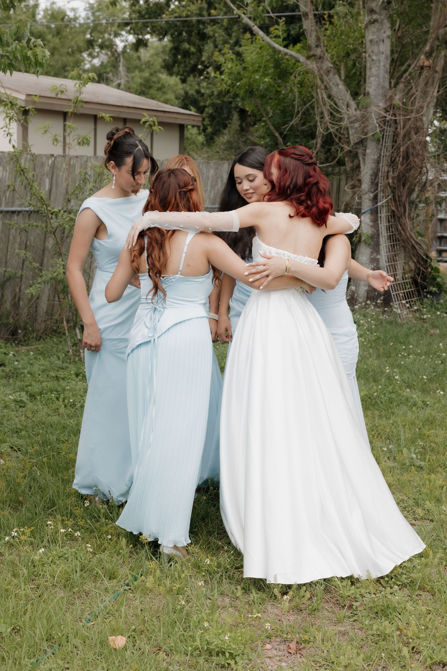 Bride in white gown hugging bridesmaids in light blue dresses outdoors in a garden setting.