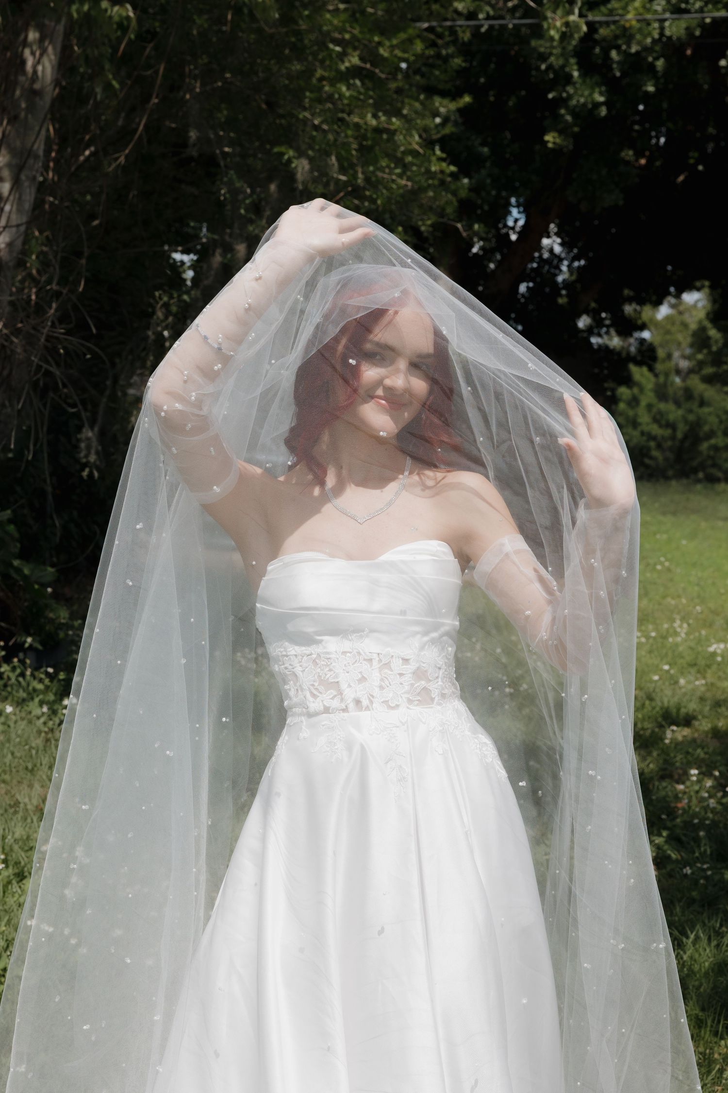 Smiling bride in strapless white wedding gown lifting her pearl-dotted veil outdoors in sunlight.