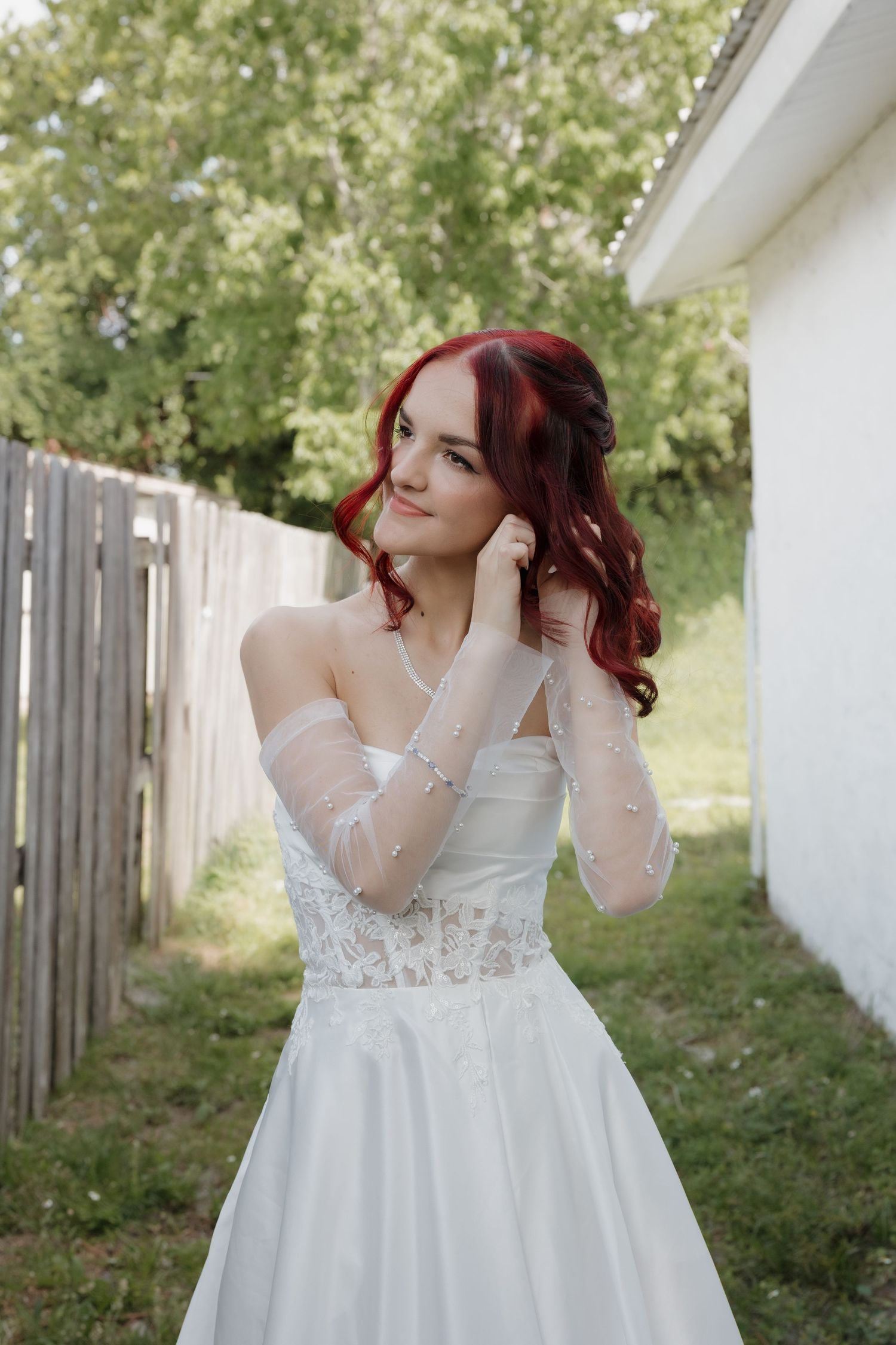 Beautiful bride with red hair in elegant off-shoulder white wedding gown posing outdoors.
