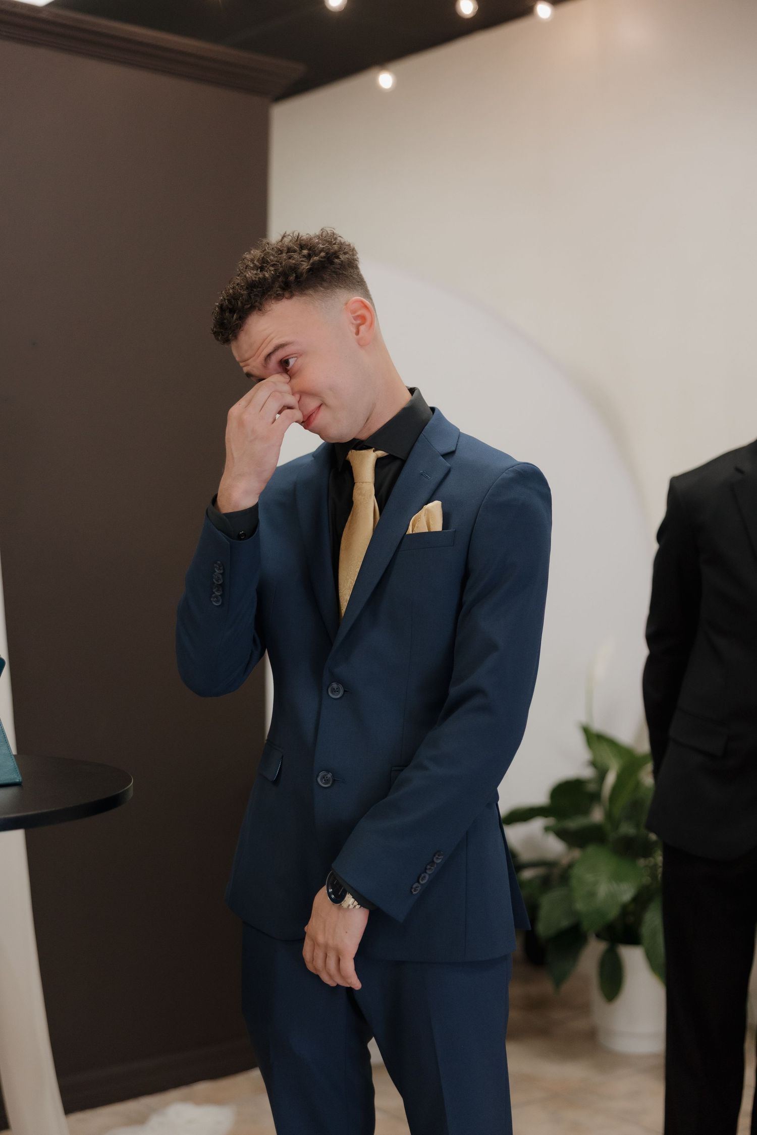 Emotional groom in navy blue suit with gold tie wiping tears before wedding ceremony.