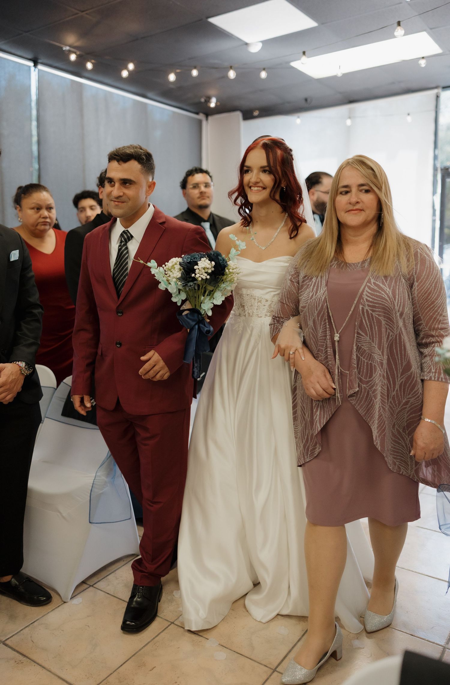 Bride in white gown with red hair walks down aisle with groom in burgundy suit.