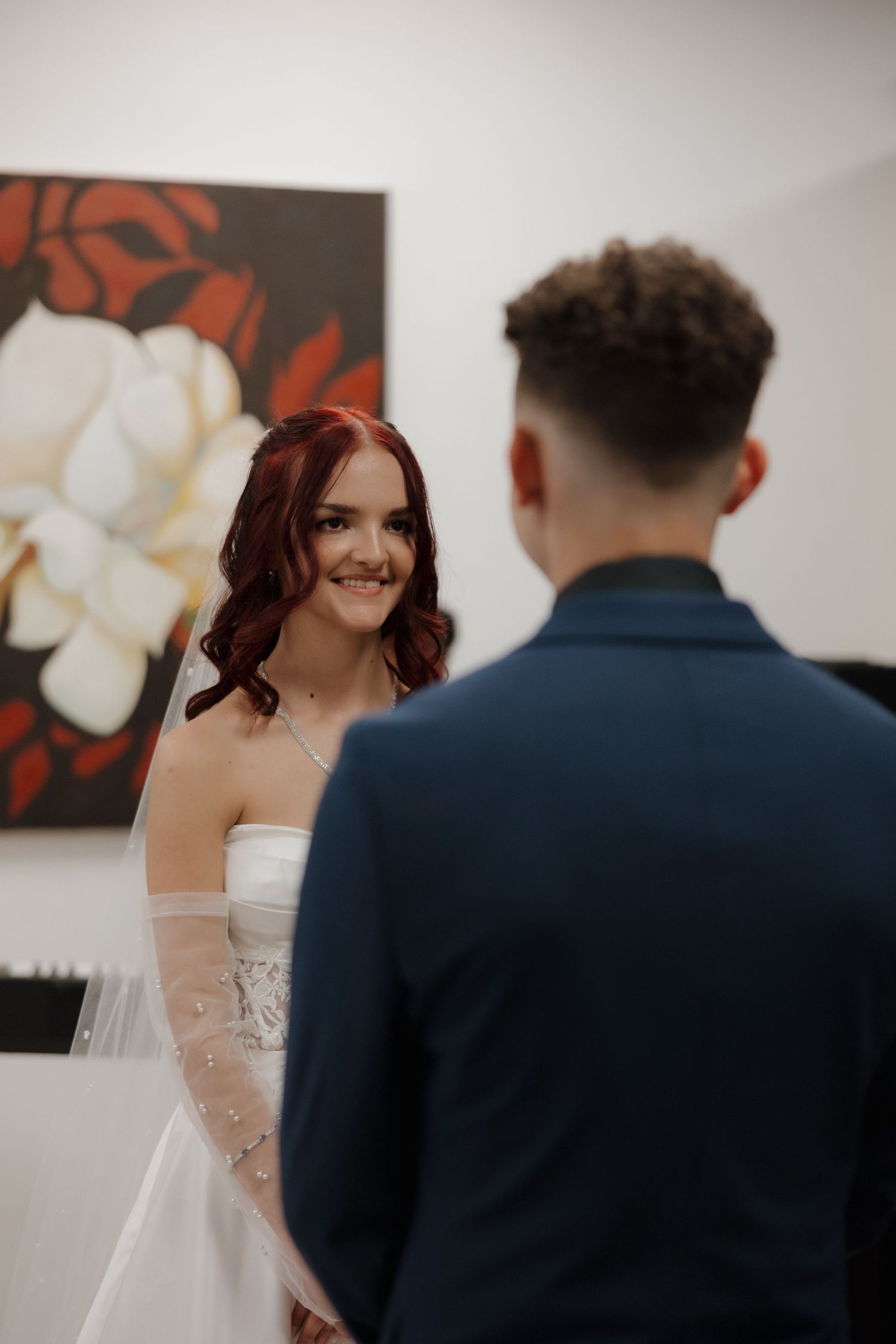Smiling bride in white gown with veil holds hands with groom in navy suit during wedding ceremony.