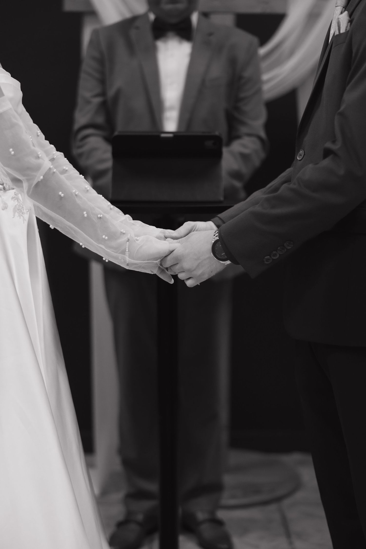 Black and white photo of bride and groom holding hands during wedding ceremony with officiant in background.