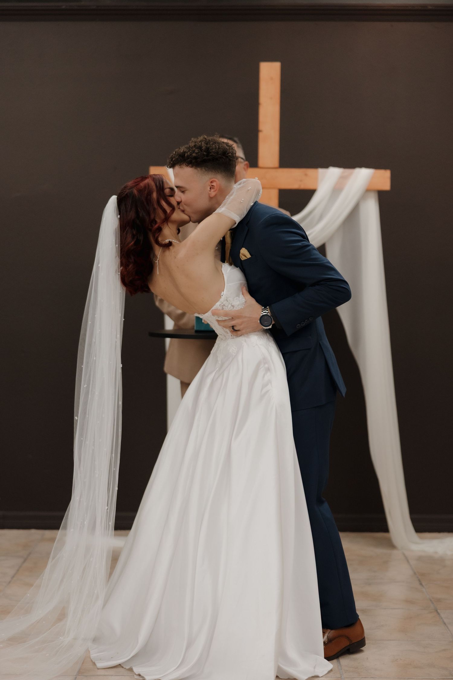 Bride and groom share first kiss at altar with wooden cross and white draping backdrop.