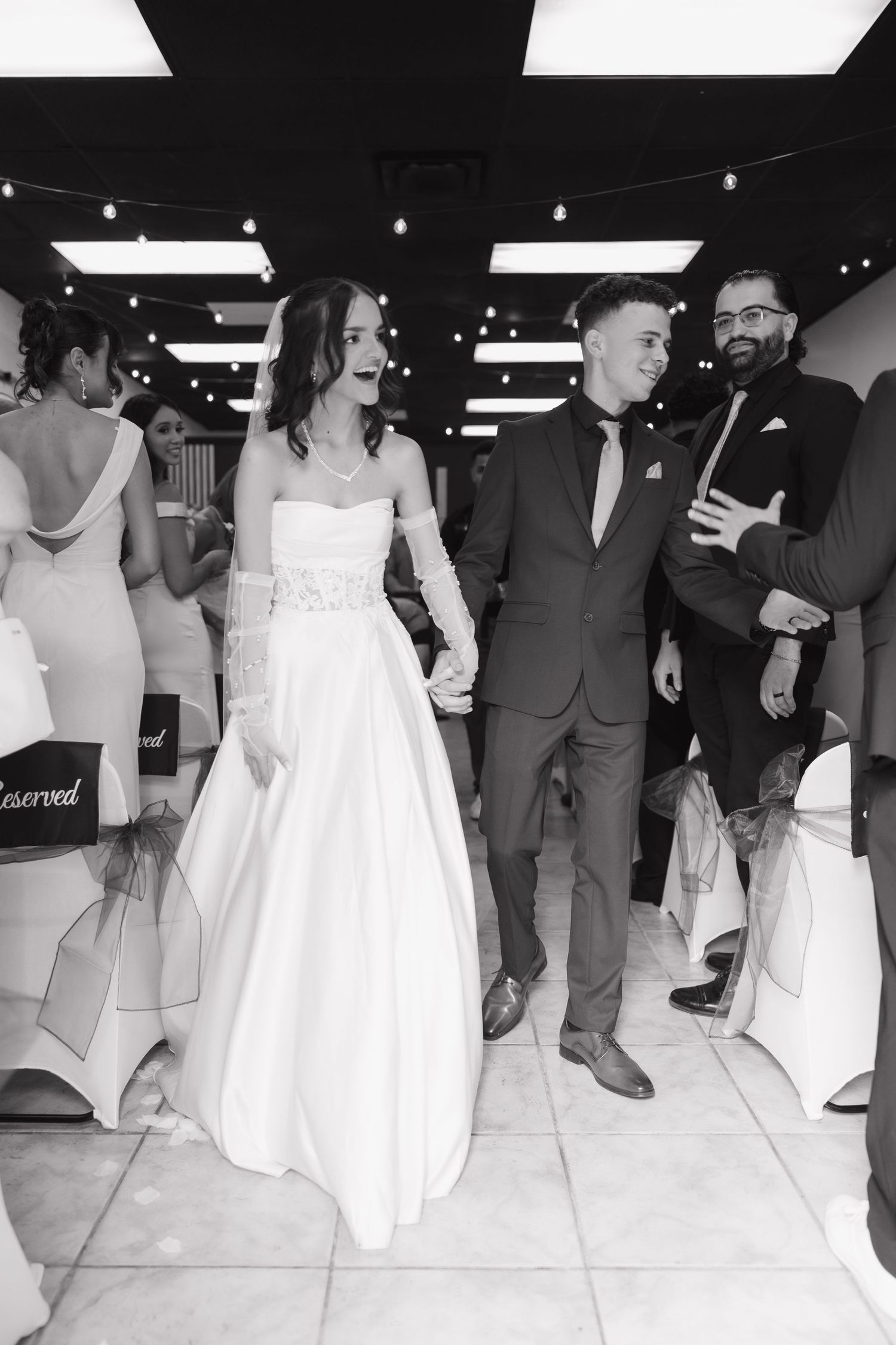 Newlyweds walk hand-in-hand down the aisle at their wedding reception in black and white photo.