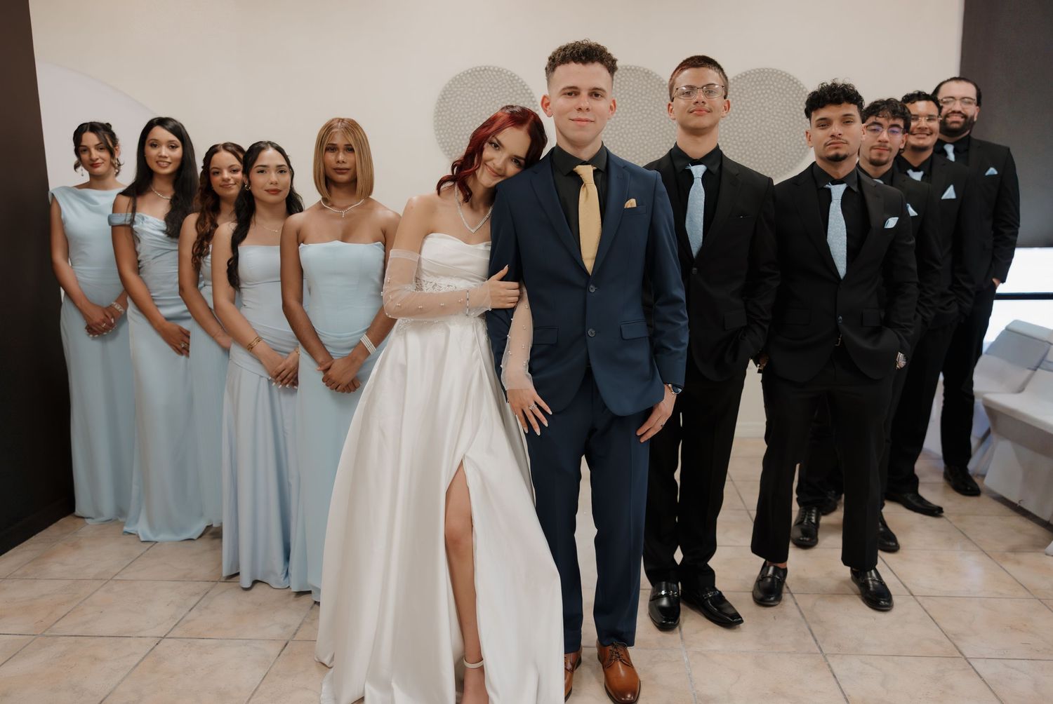 Wedding party poses in light blue bridesmaid dresses and navy suits, with bride in white gown and groom in navy tuxedo.