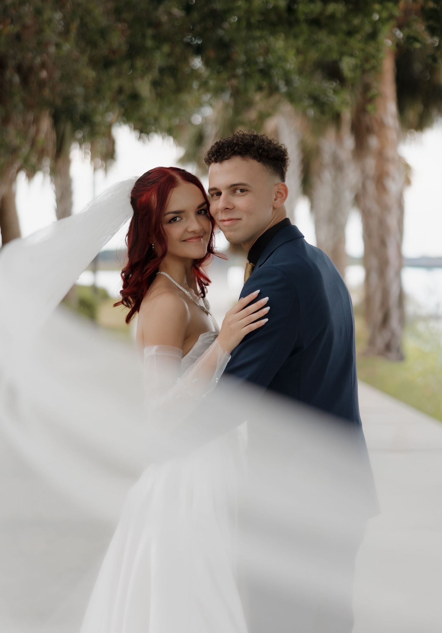 Bride in white gown with red hair and groom in navy suit pose outdoors with veil flowing.