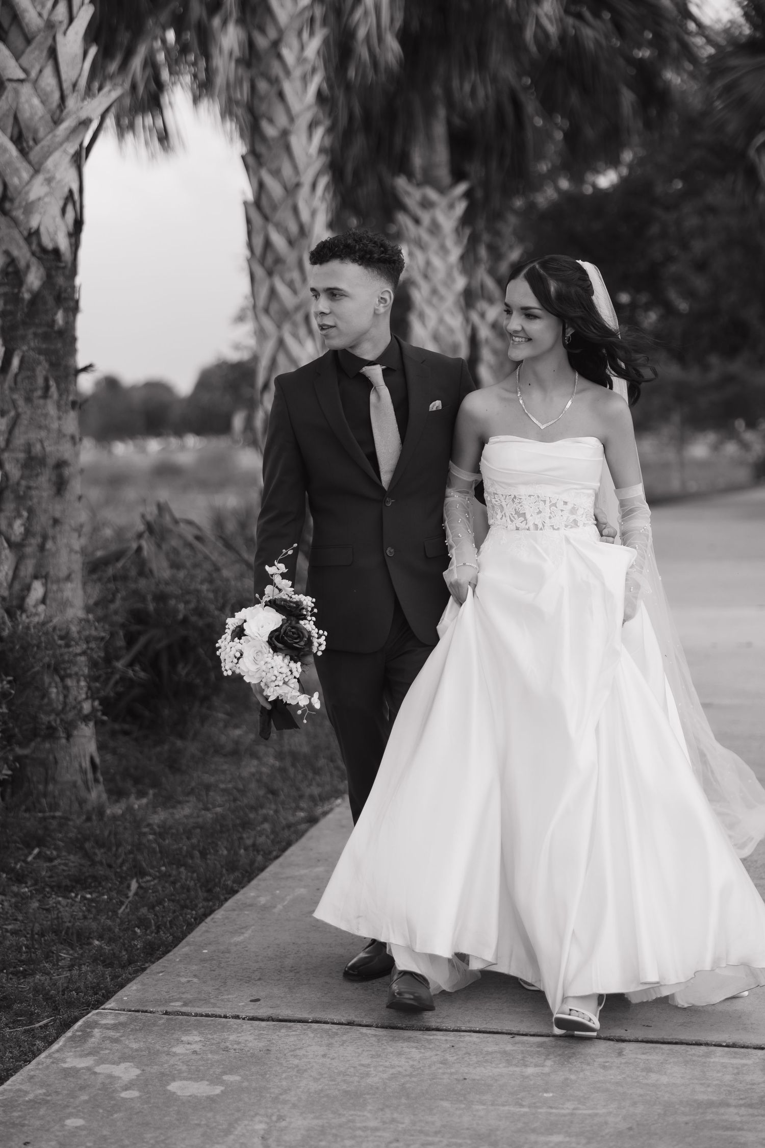 Black and white photo of a bride and groom walking together outdoors near palm trees on their wedding day.