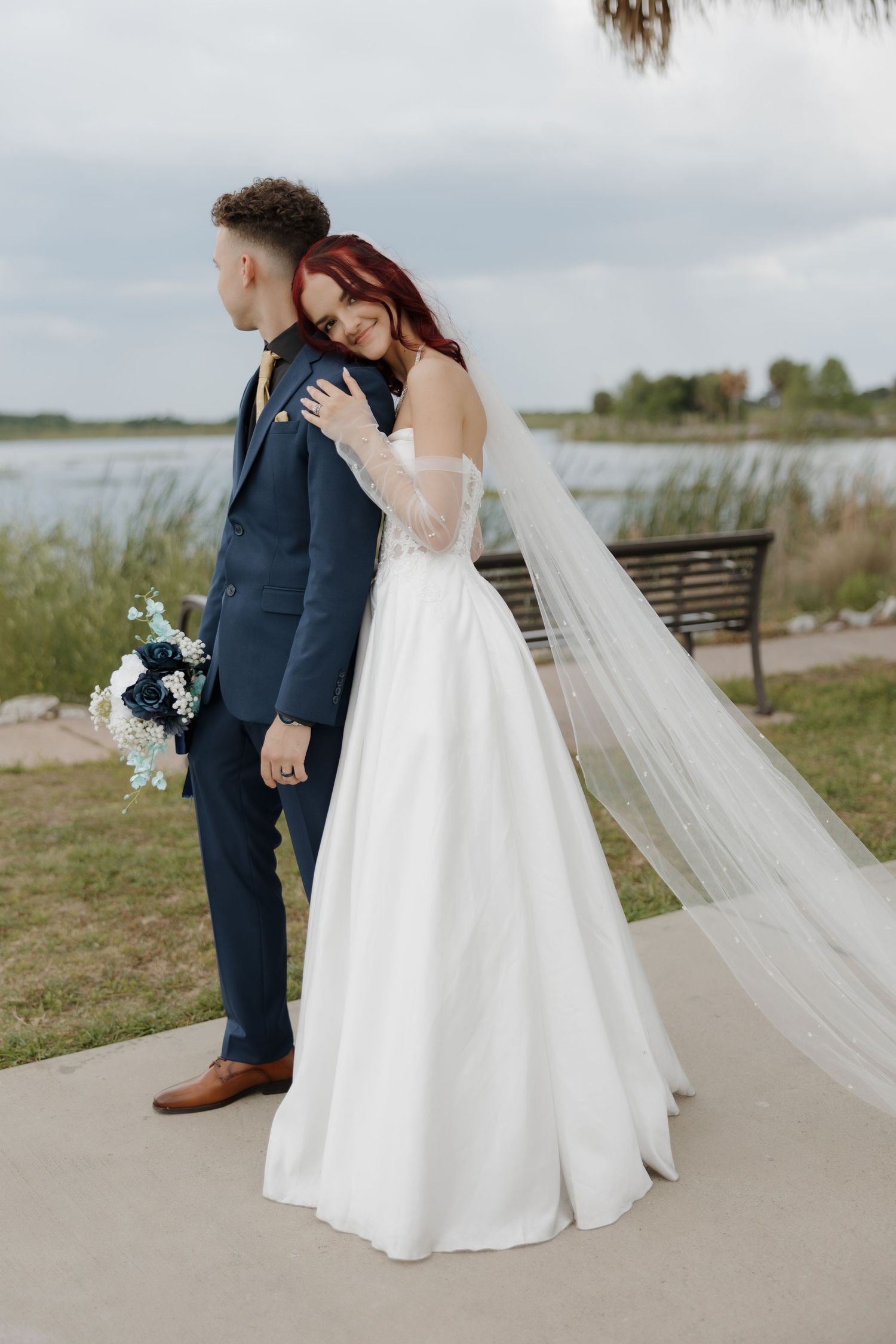 A bride with red hair in a white gown embraces her groom near a scenic lakeside on their wedding day.