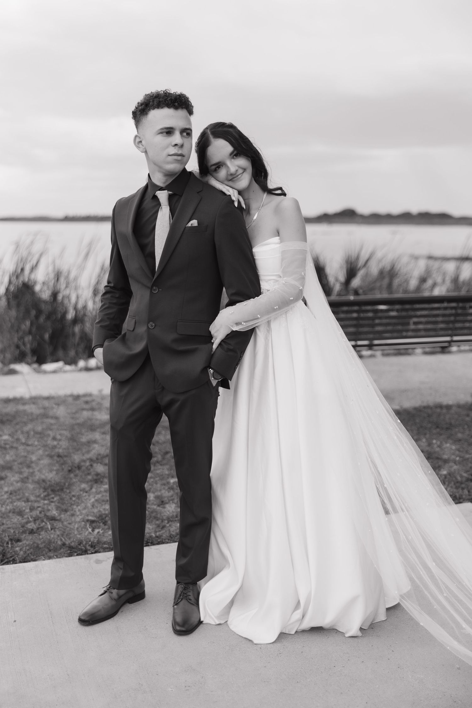 Black and white portrait of bride leaning on groom in dark suit near waterfront venue.