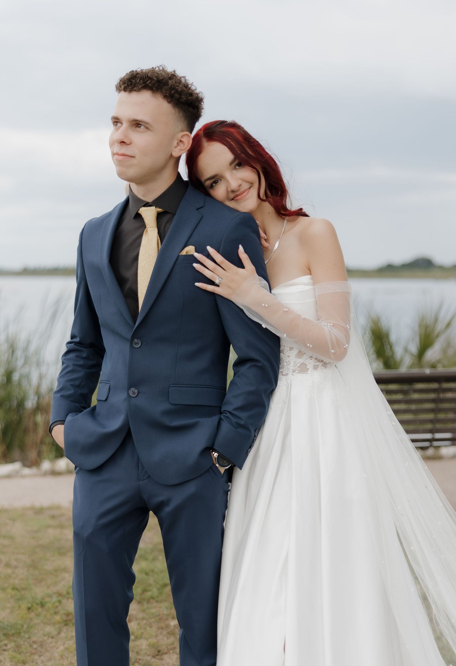 Wedding couple posing outdoors by lake, groom in navy suit with gold tie, bride in white gown.