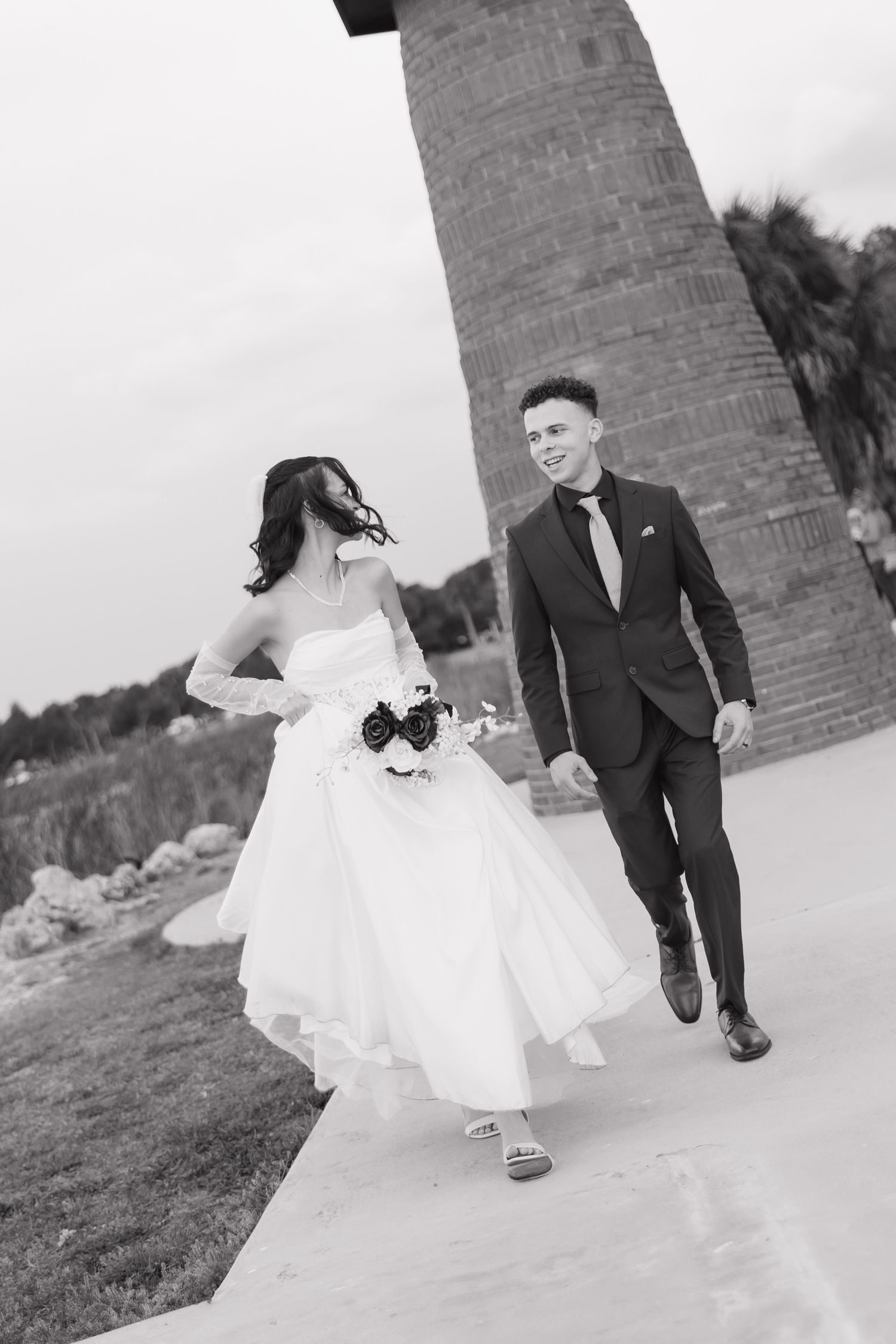 Black and white photo of bride and groom walking together near brick lighthouse outdoors.