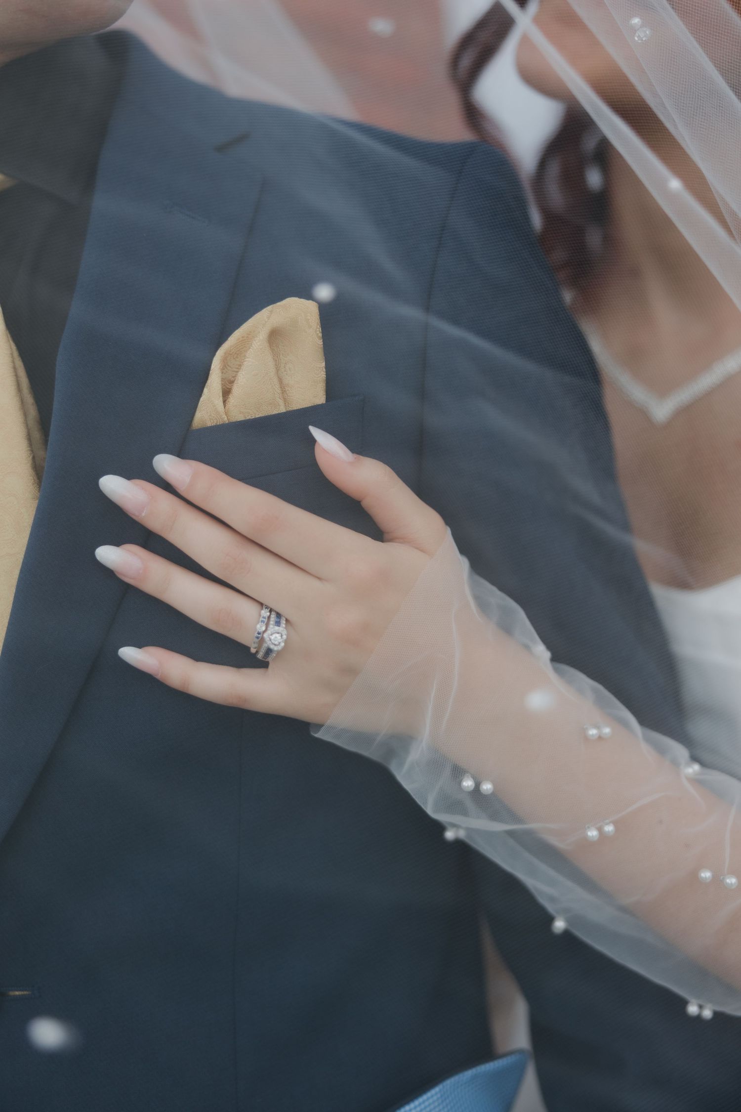 Bride's hand with diamond ring and pearl-adorned veil resting on groom's navy suit with gold pocket square.