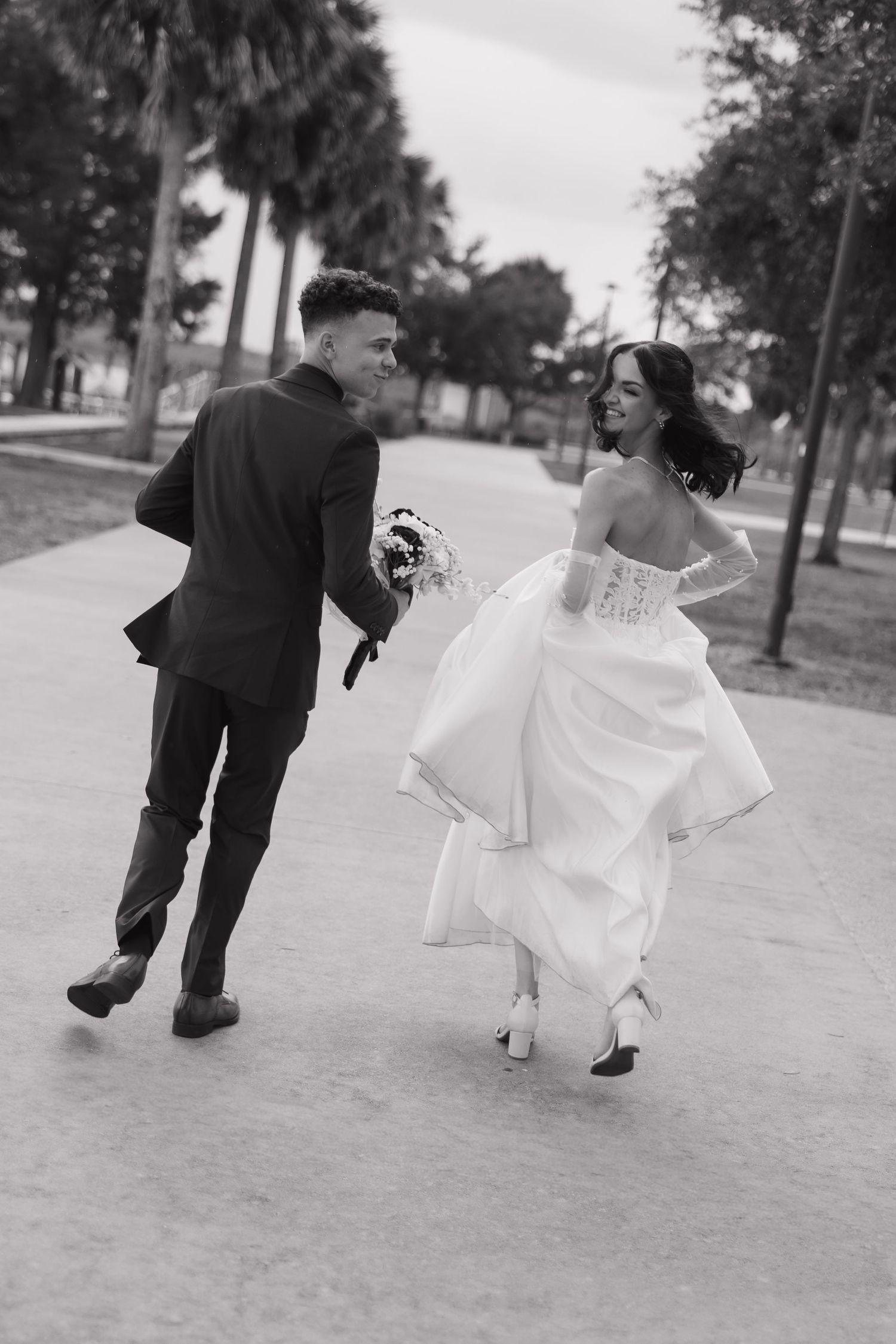 Black and white photo of newlywed couple joyfully walking together outdoors with bouquet on wedding day.