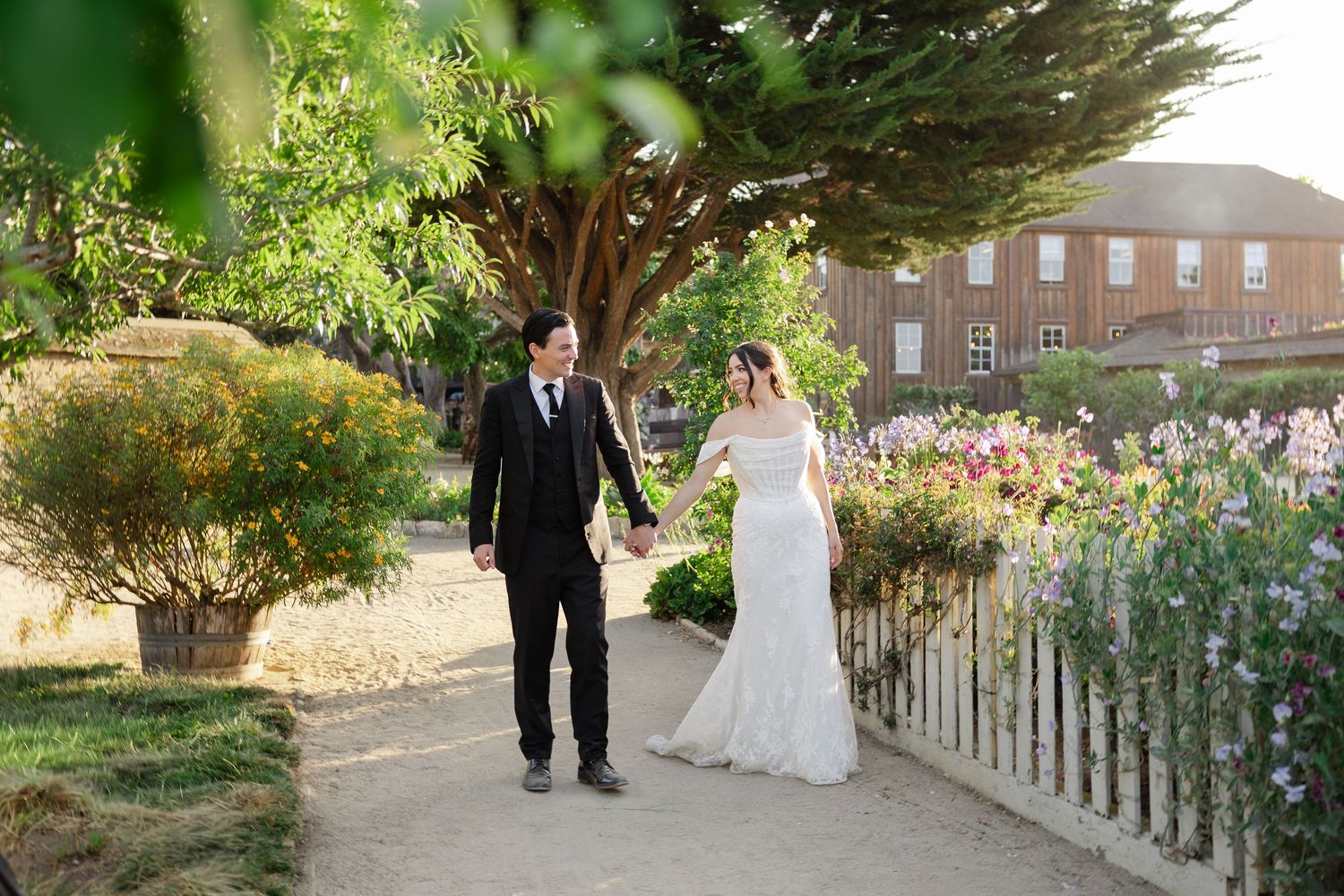Bride and groom walk hand-in-hand along a garden path lined with colorful flowers and lush trees on their wedding day.