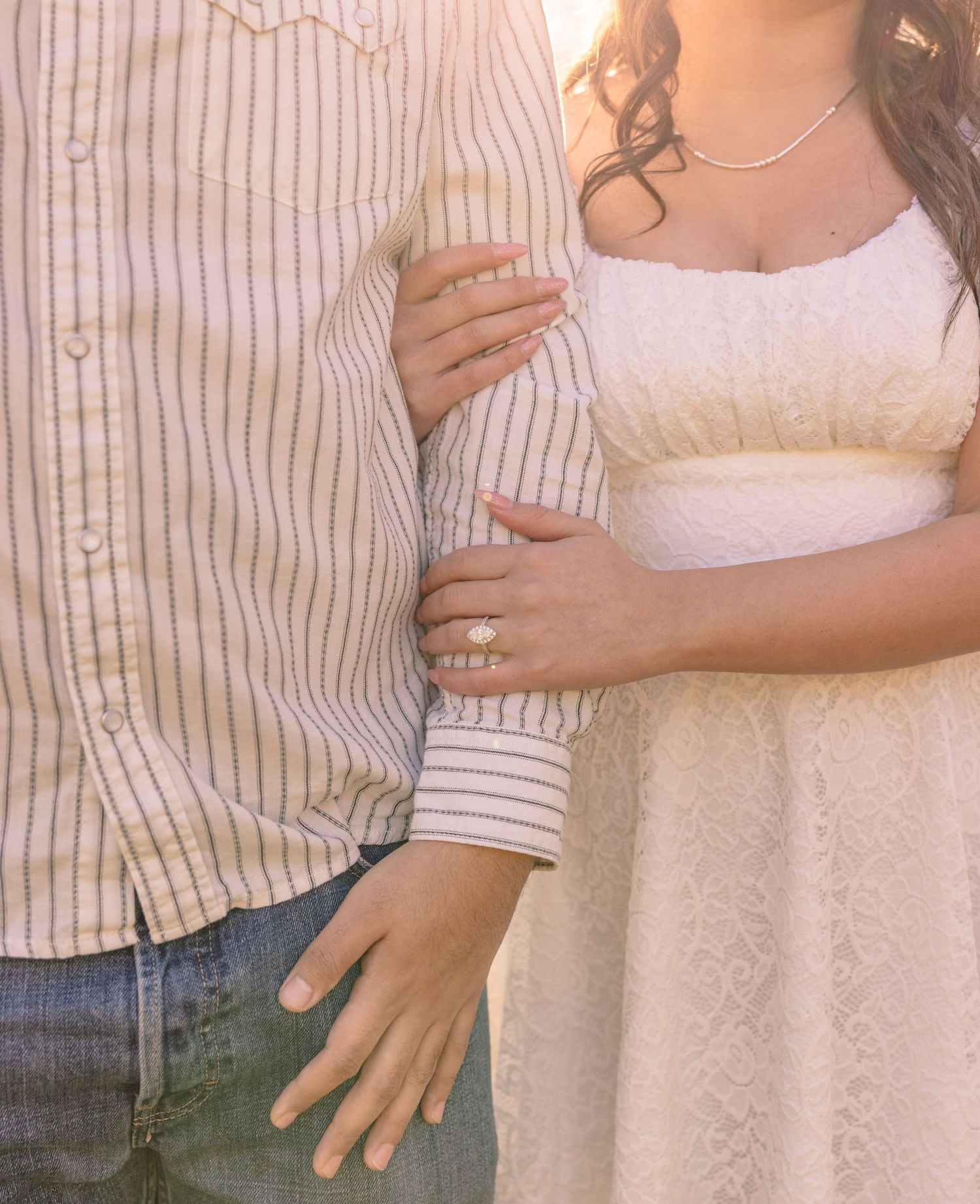 Couple embracing, woman wearing engagement ring and white lace dress, man in striped shirt and jeans.
