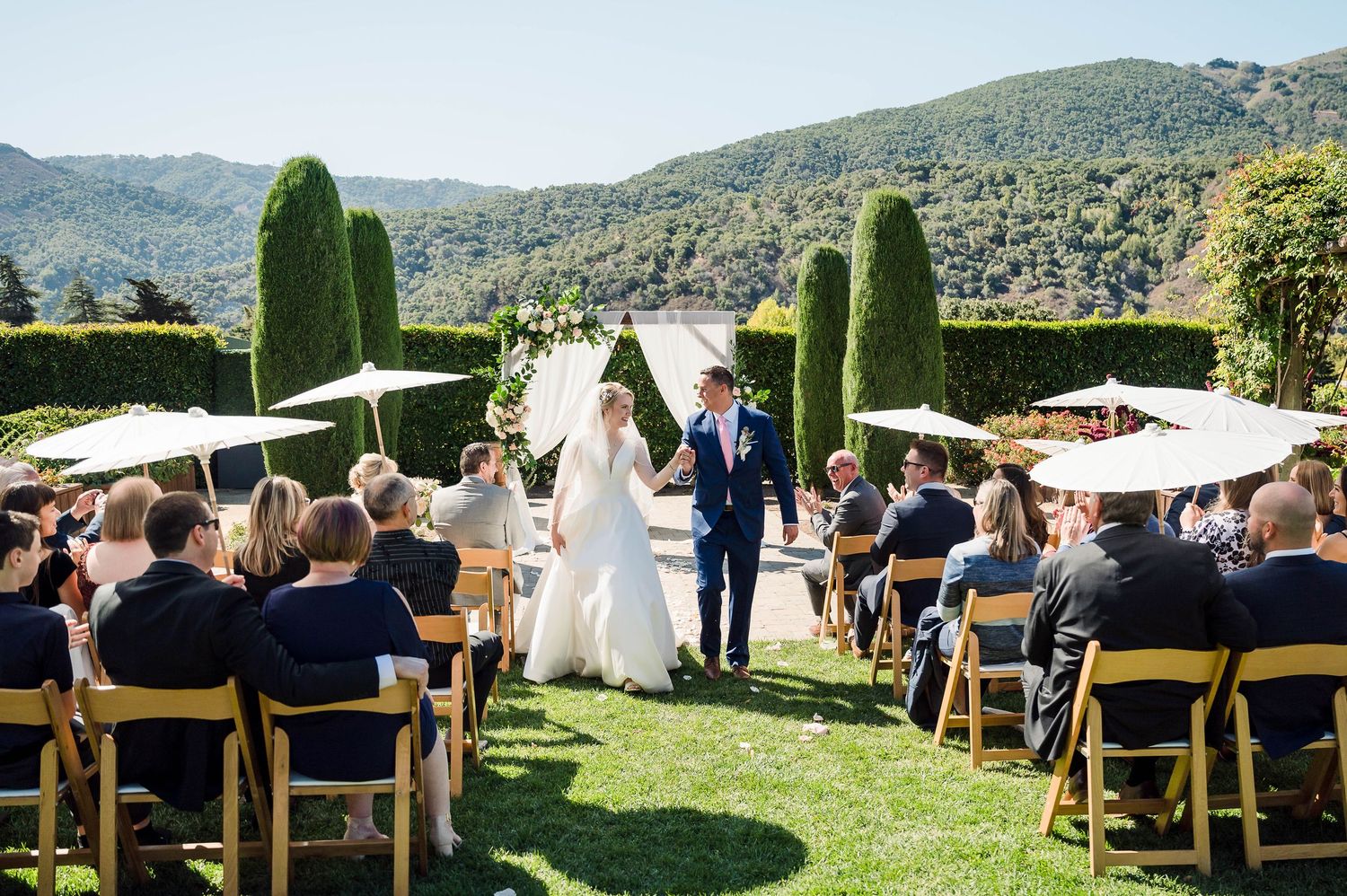 People gather under a large tree for an outdoor celebration with wooden benches and decorative lighting.