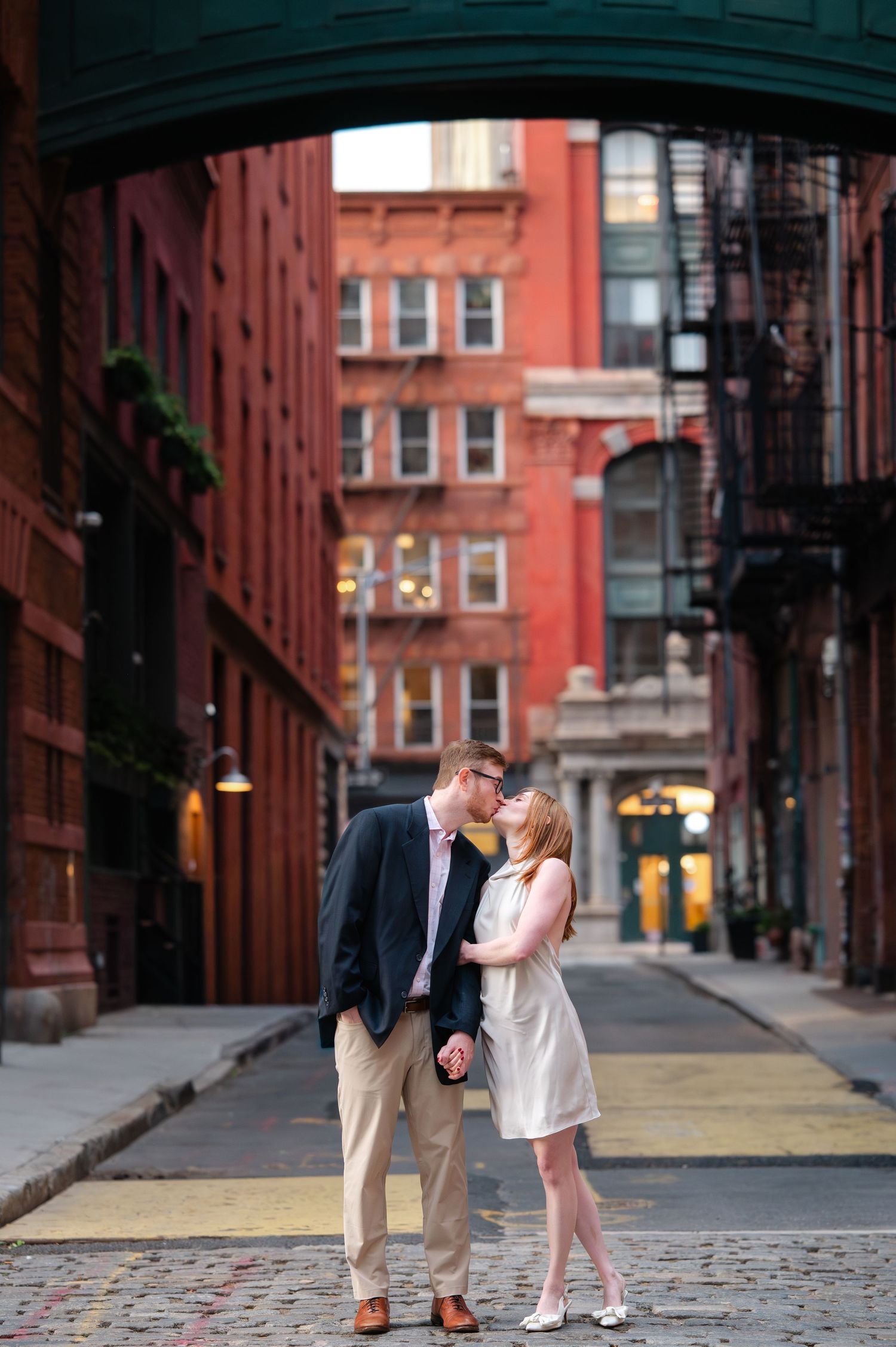Couple sharing a romantic kiss on cobblestone street in Tribeca, NYC, surrounded by red brick buildings during their engagement session.