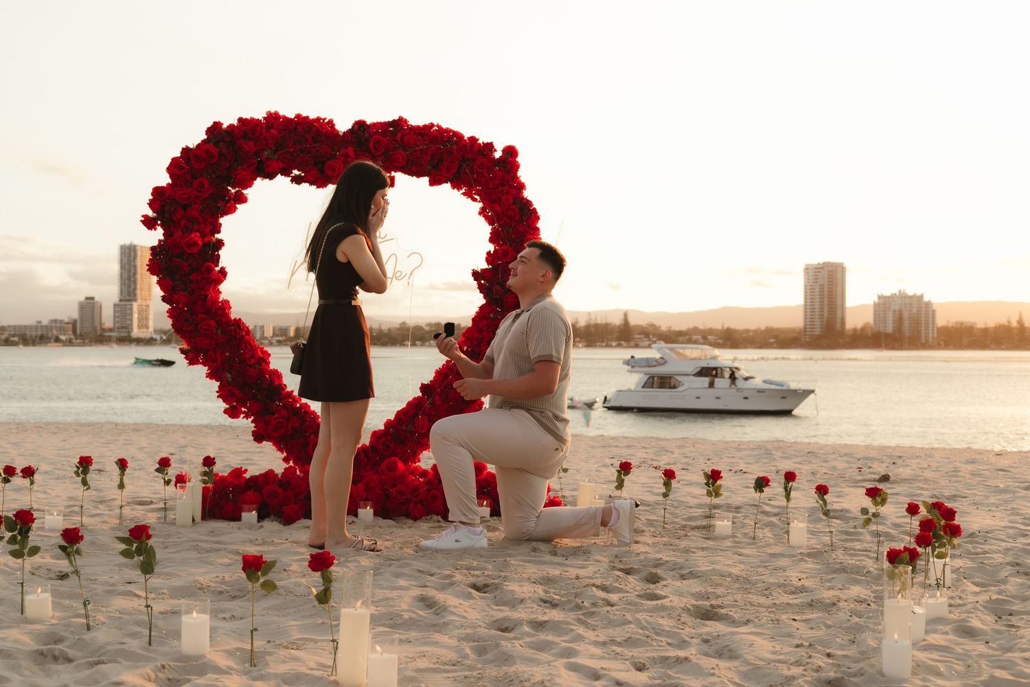 Man proposes to woman on beach surrounded by red roses and heart-shaped floral arch at sunset.