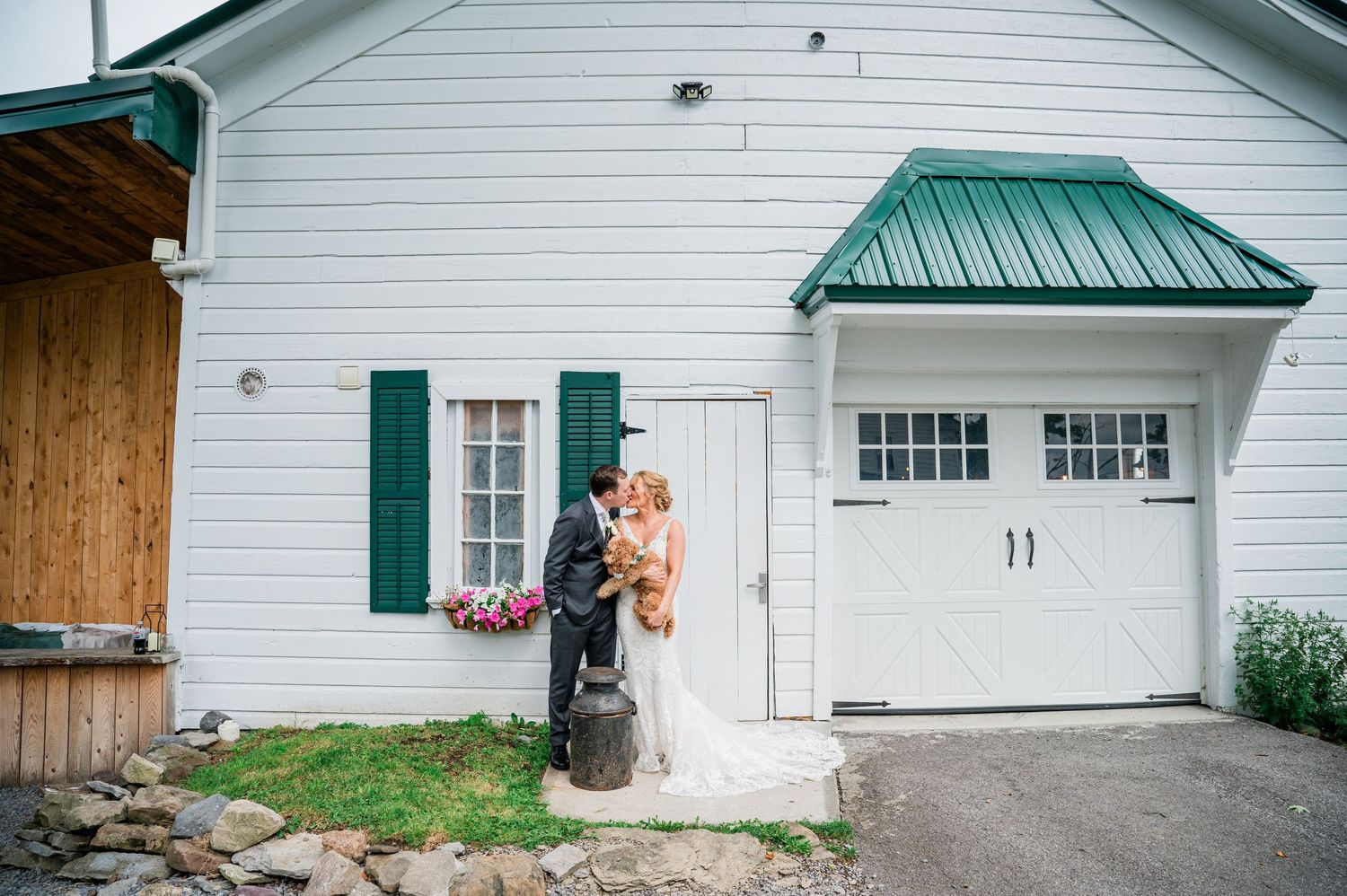 Bride and groom sharing a kiss outside a white barn wedding venue with green shutters and metal roof at Knotting Hill Farm Jordanville, NY.