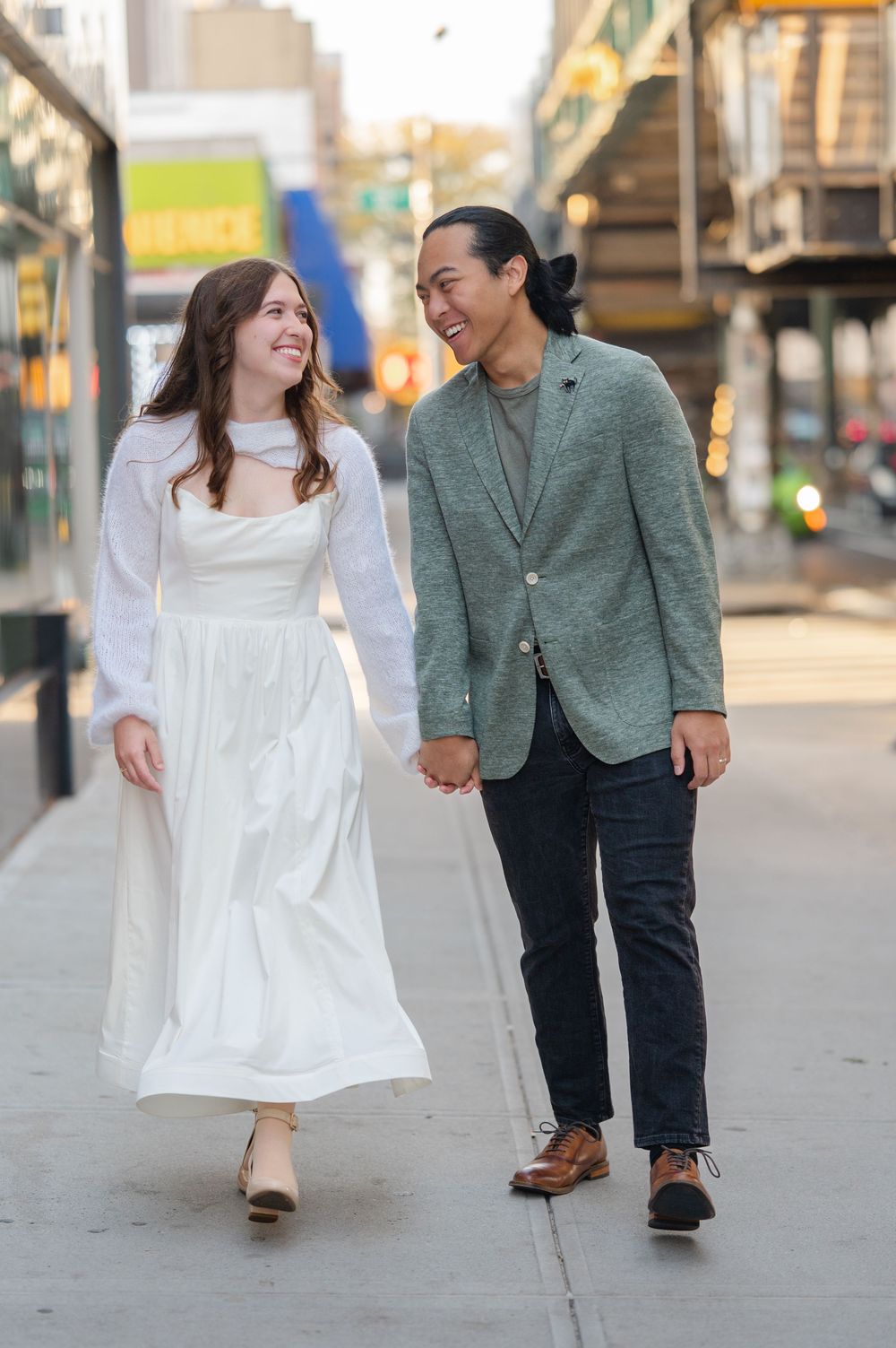 Smiling couple walks hand-in-hand on urban sidewalk, woman in white dress and man in green blazer.