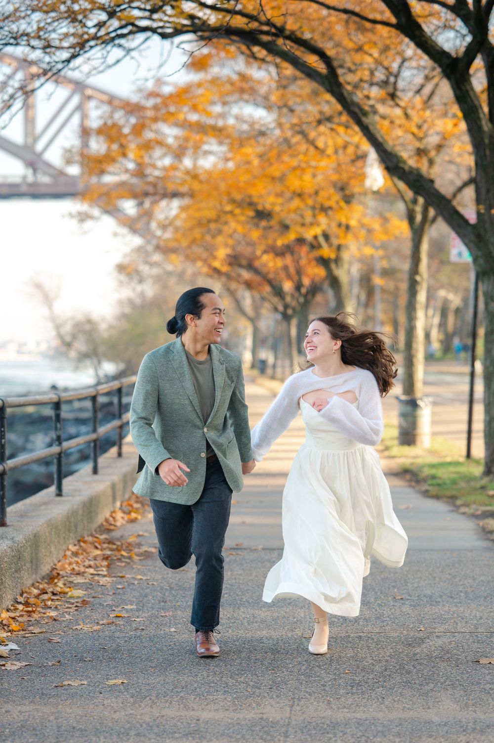 Joyful couple running hand in hand along autumn tree-lined path near bridge, woman in white dress laughing.