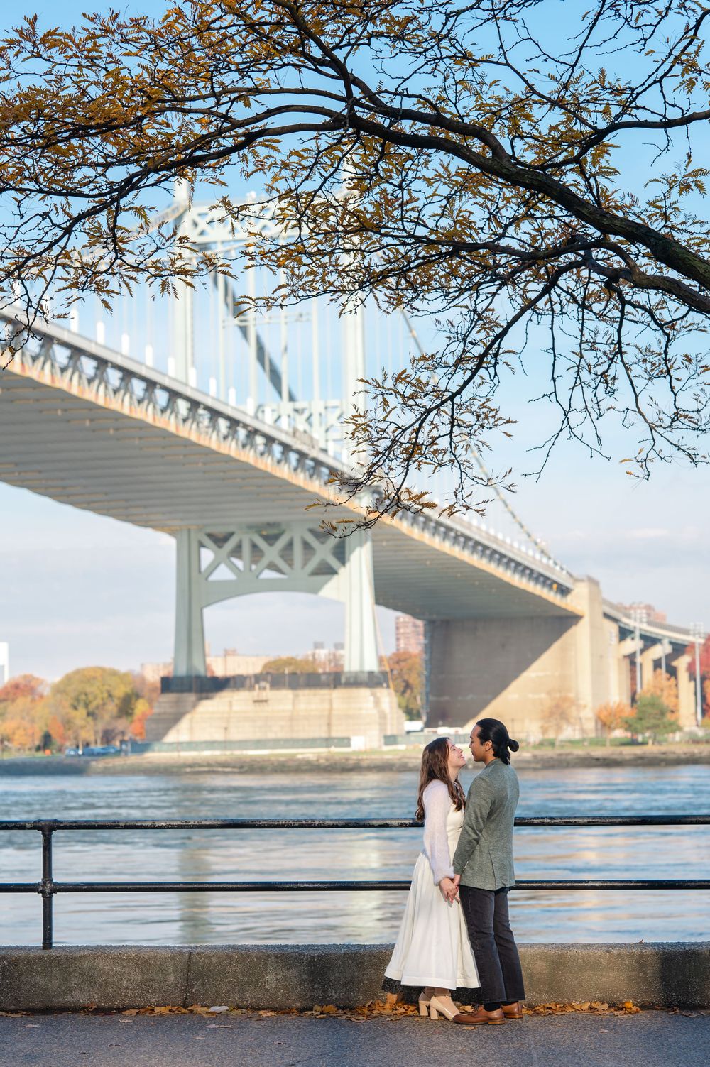 Couple holds hands facing each other beneath a large suspension bridge by the river.