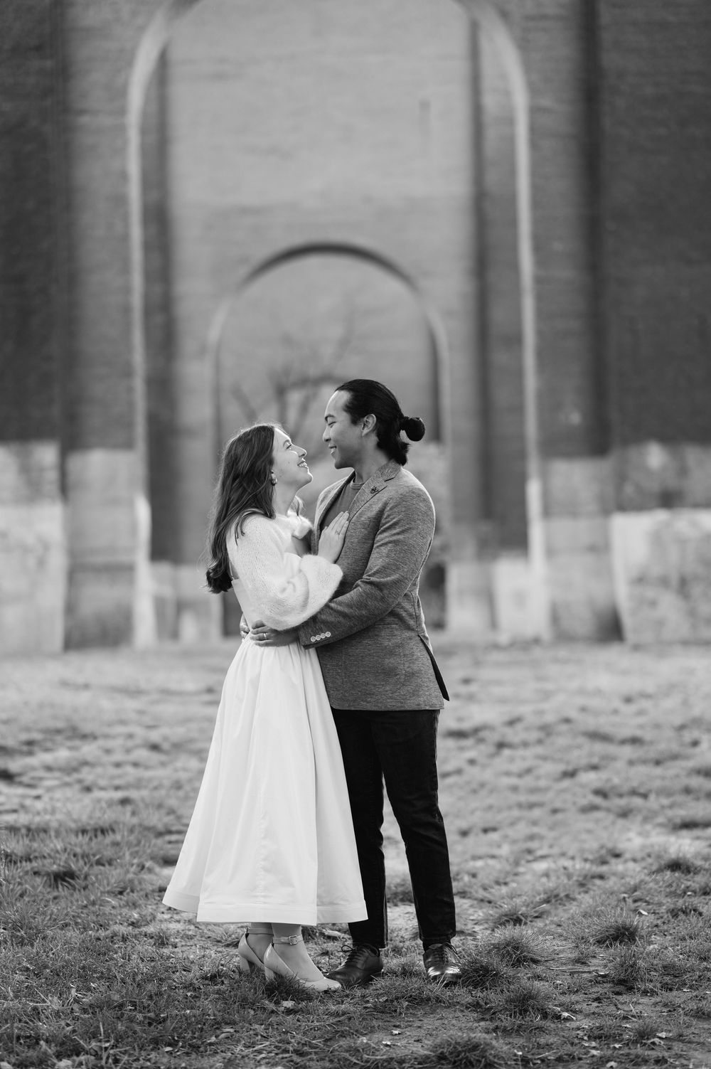 Black and white photo of couple about to kiss near brick archway.