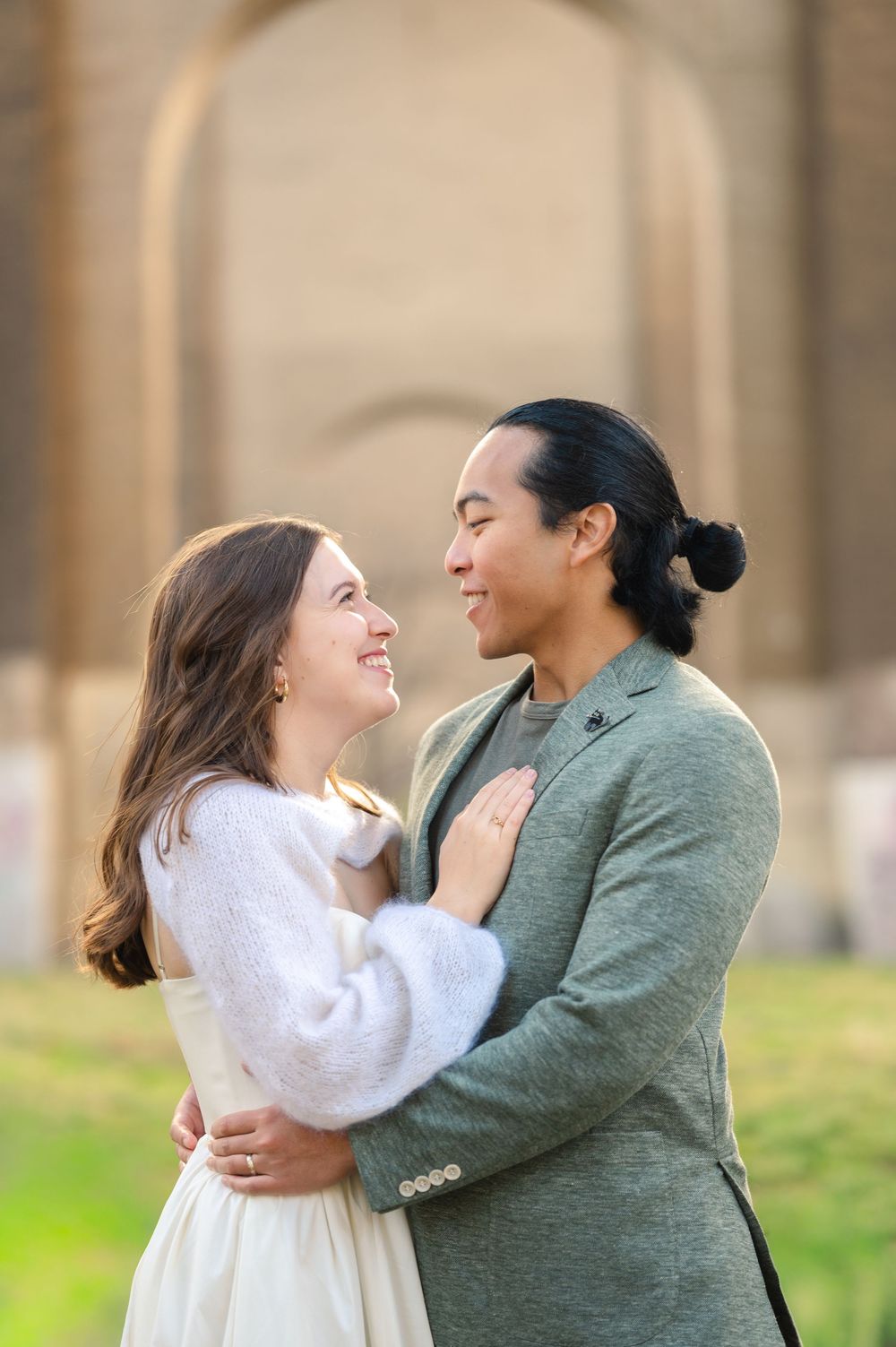 Smiling couple embracing outdoors near an arched stone structure, woman in white dress and man in green blazer.
