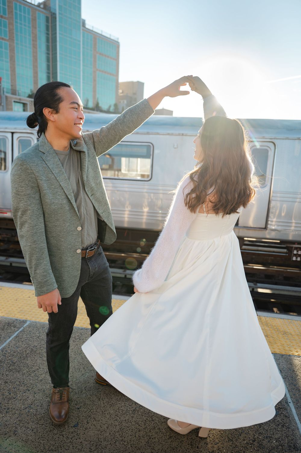 Couple dancing on a train platform in golden sunlight, man in gray blazer spinning woman in flowing white dress.