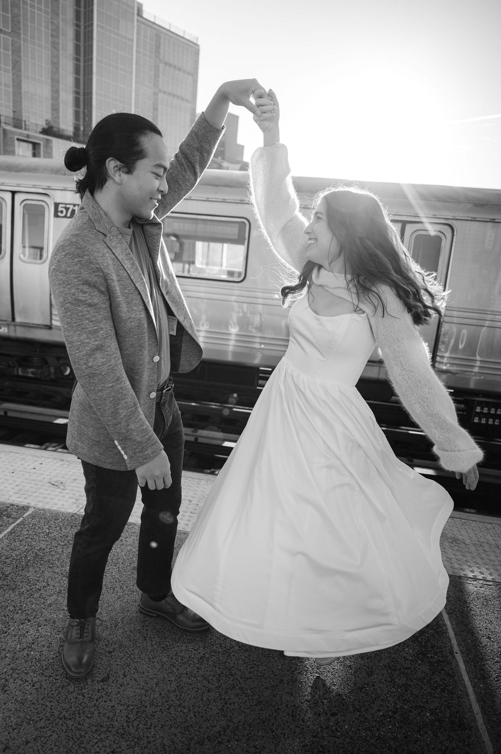 Black and white photo of couple dancing on sunny subway platform, woman in white dress twirling joyfully.