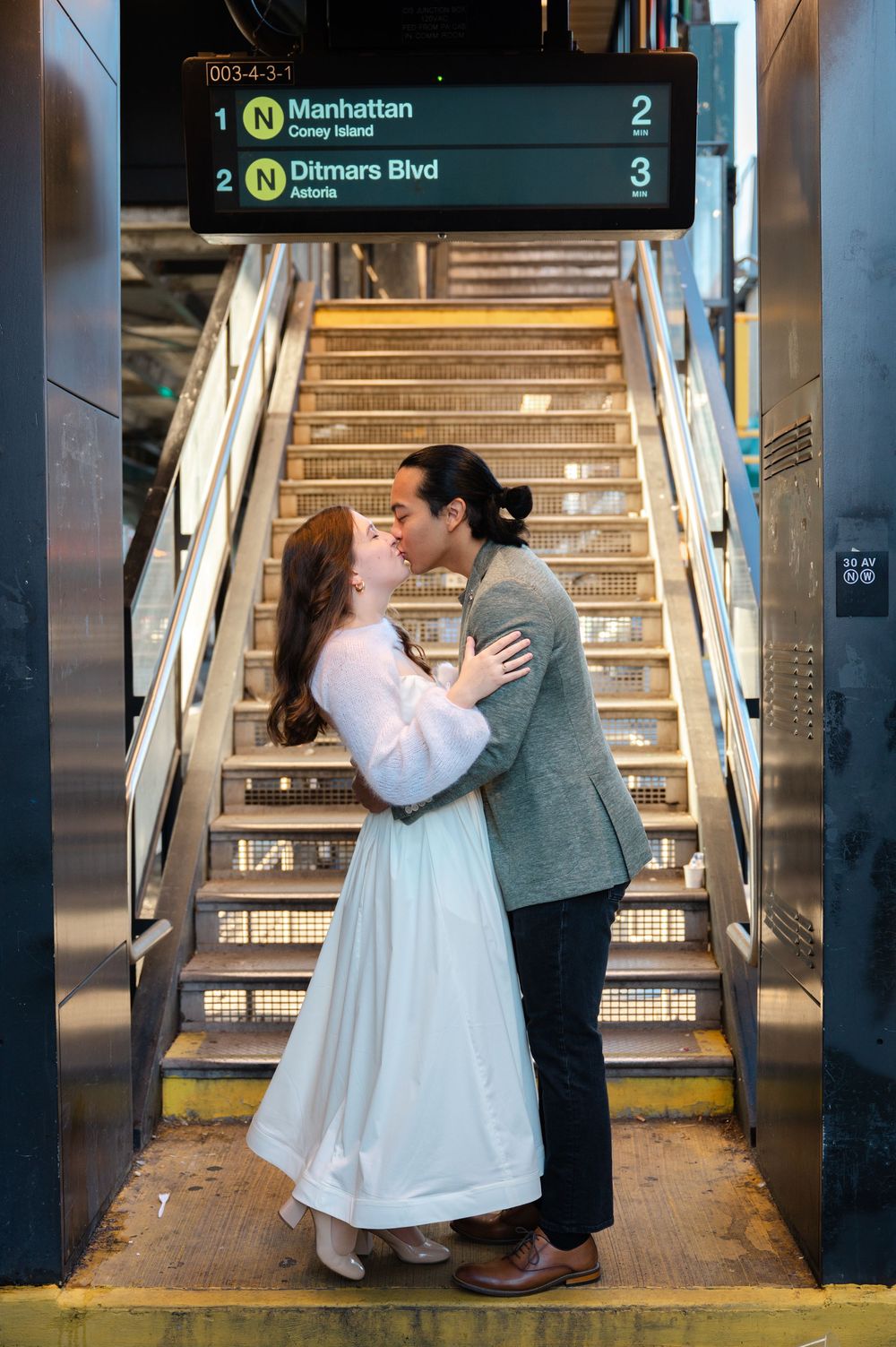 Couple kissing on subway stairs under Manhattan N train sign, woman in white dress embracing man in gray blazer.