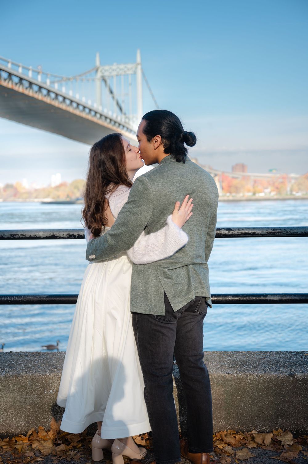 Couple sharing a kiss by waterfront railing with large suspension bridge and blue sky in background.