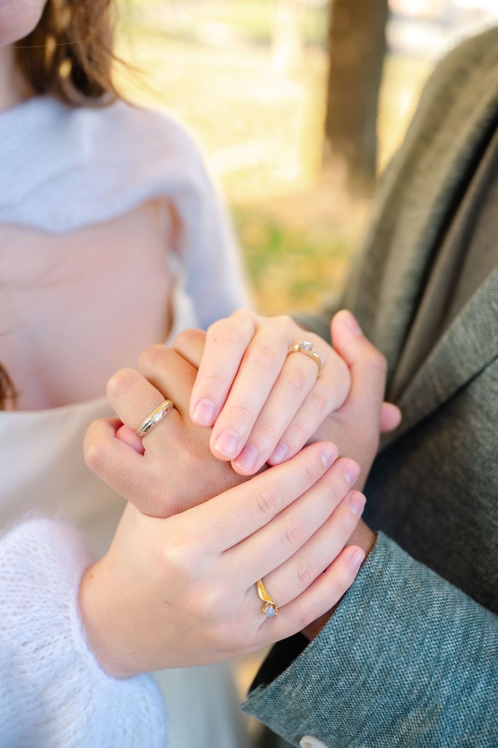Close-up of couple holding hands displaying gold engagement and wedding rings in an outdoor setting.