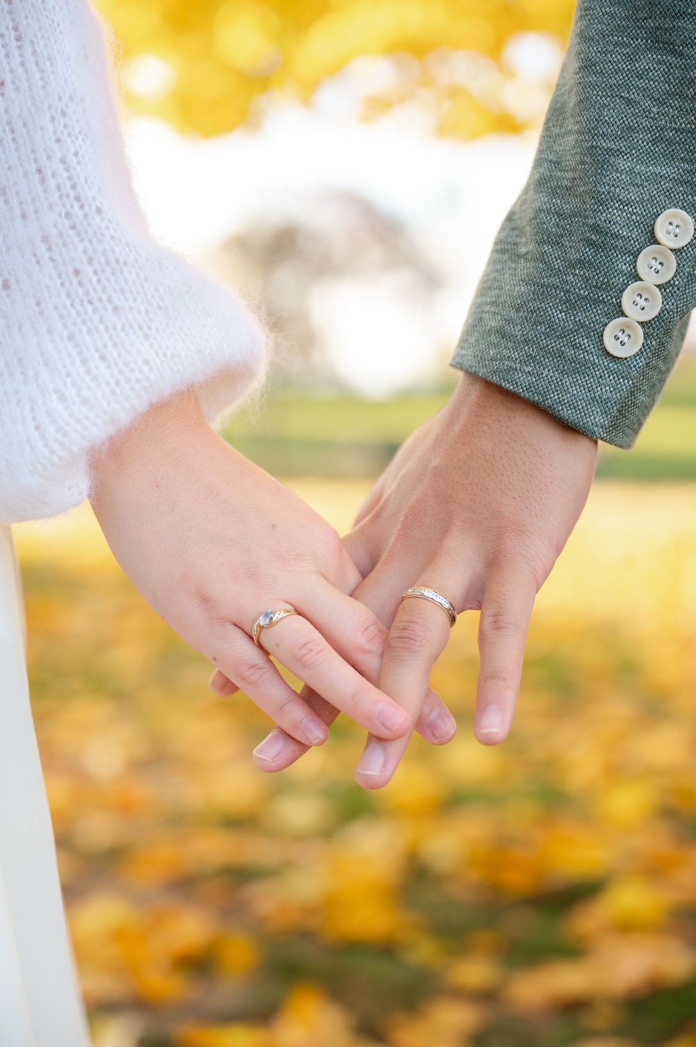 Couple holding hands showing wedding and engagement rings with autumn leaves in background.