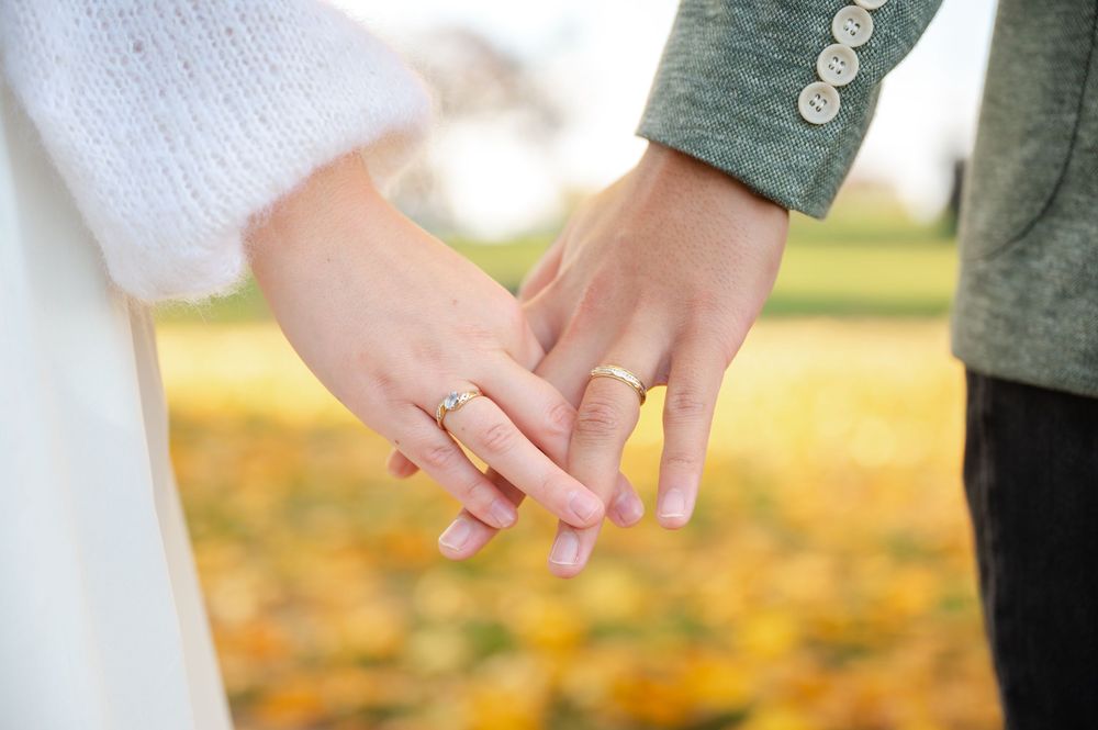 Close-up of engaged couple holding hands showing diamond engagement ring and wedding band in autumn park.