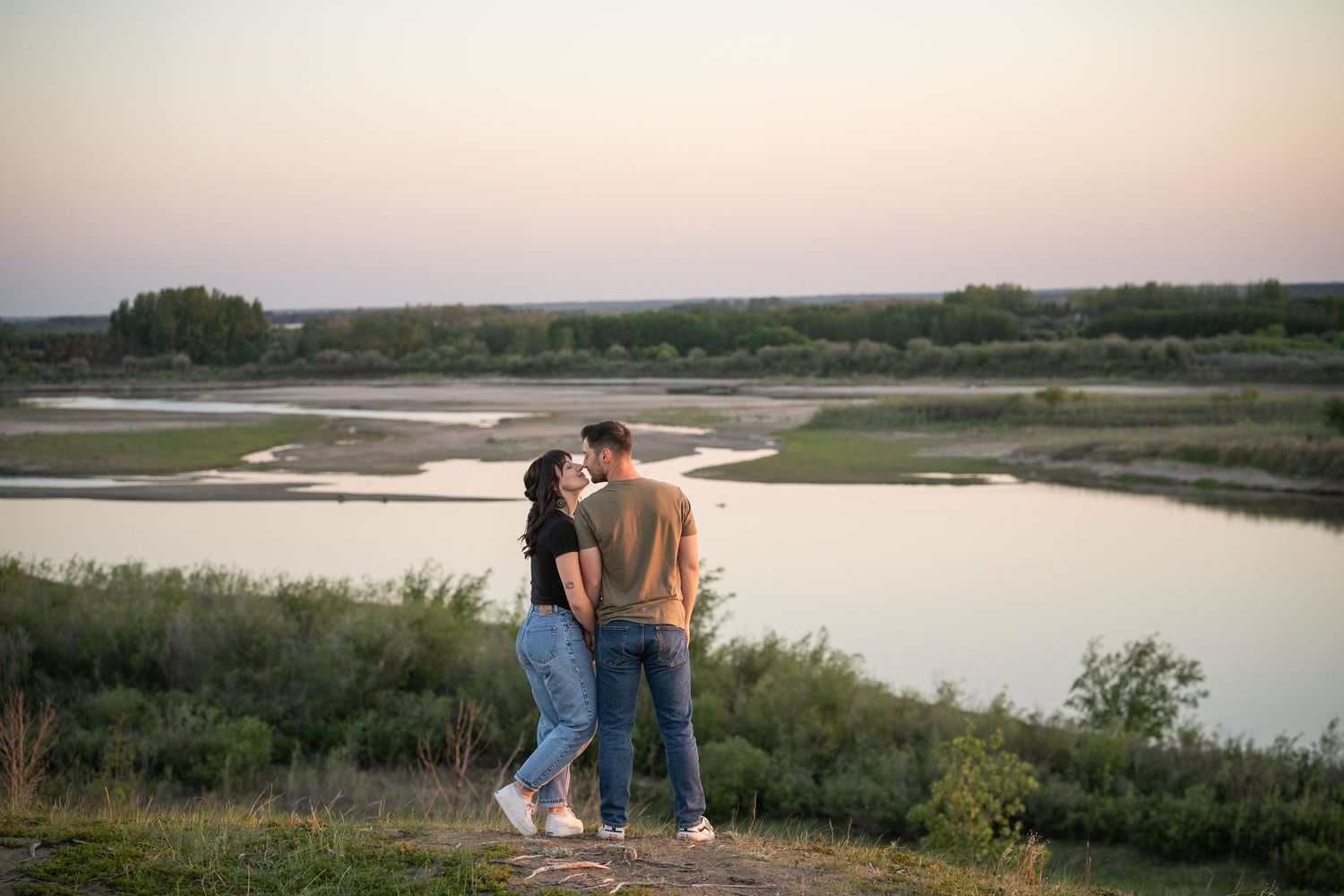 Couple sharing a romantic kiss at sunset overlooking a scenic river landscape with lush greenery.