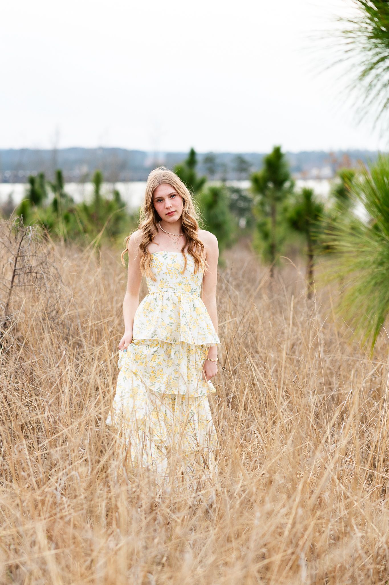 A smiling teenage girl in a yellow floral sundress stands in a field of dry grass with evergreen trees behind her.