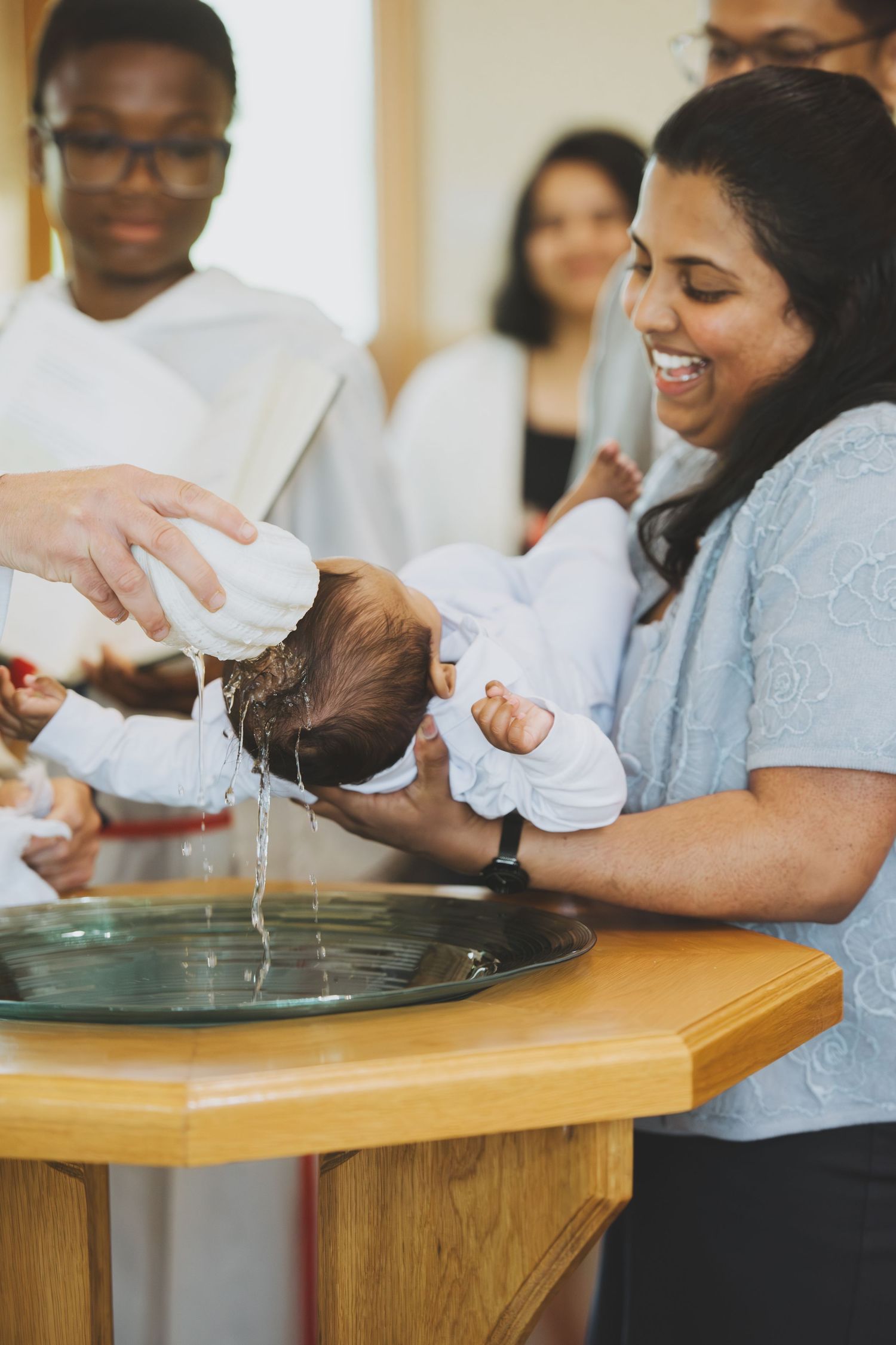 A smiling woman holds an infant during a Christian baptism ceremony at a church font.