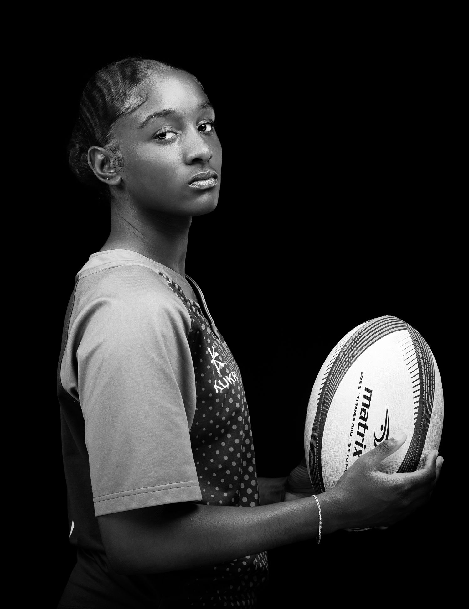 Black and white image of a female rugby player holding a rugby ball, looking at camera from the side