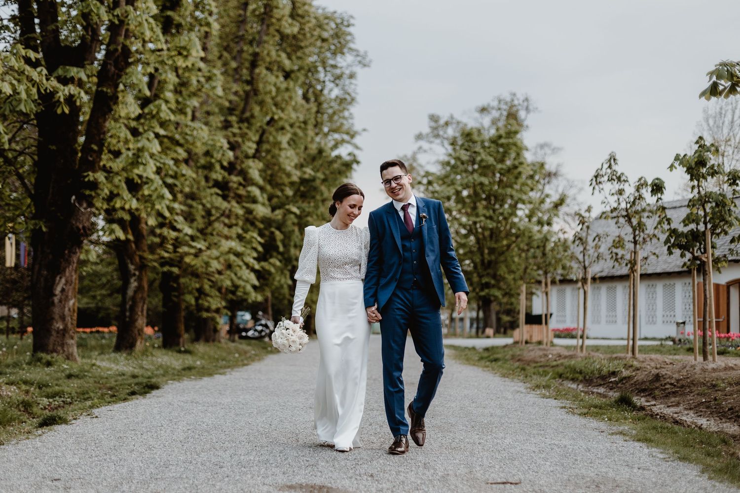 Newlywed couple walking together on tree-lined gravel path.
