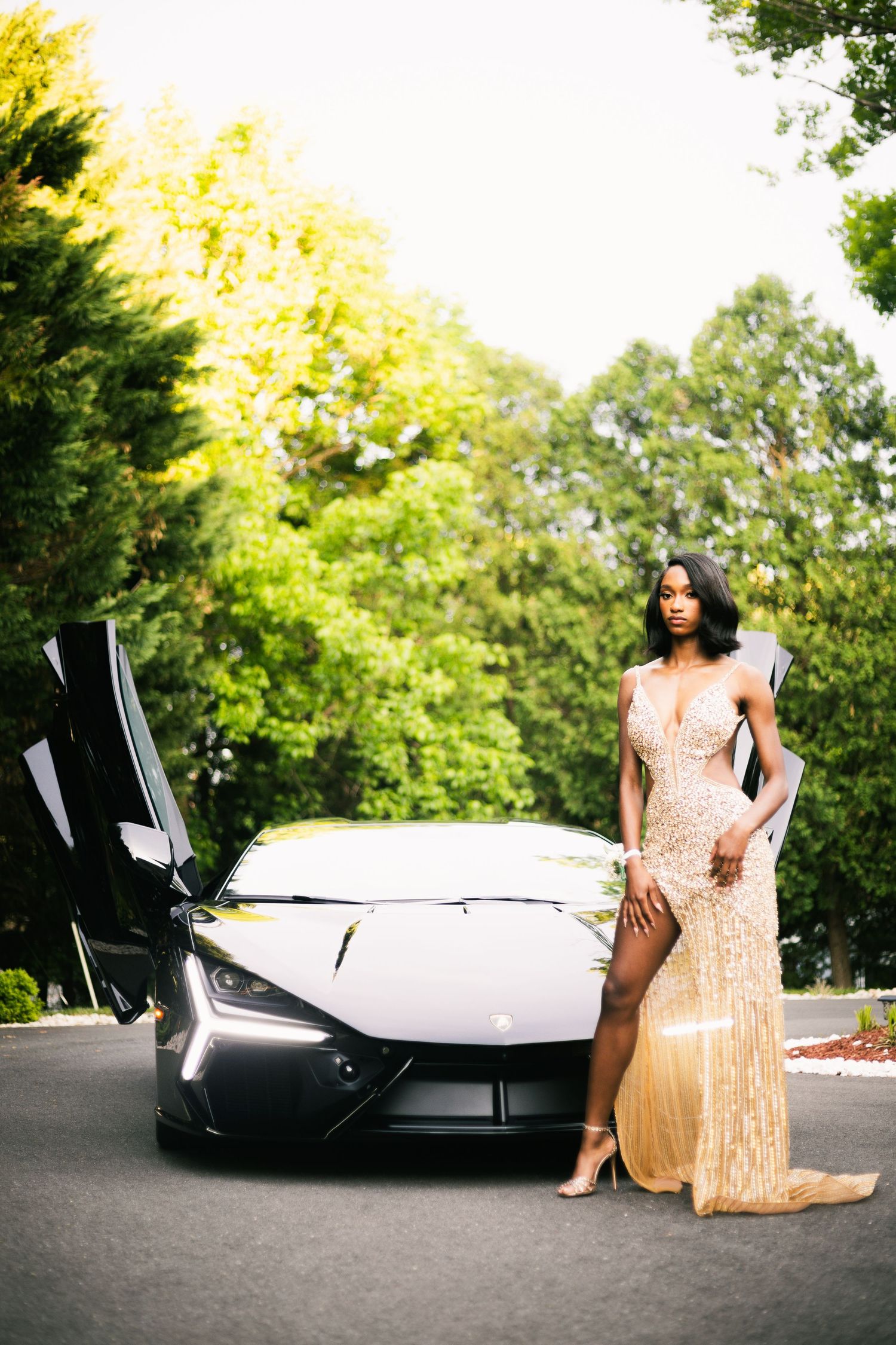 A woman in a gold sequin gown poses elegantly beside a black Lamborghini with scissor doors open outdoors.