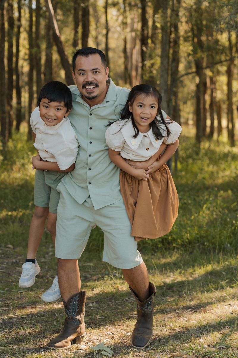 Smiling father playfully holds his young children in a sunny forest setting, all dressed in casual, coordinated outfits. Family photoshoot