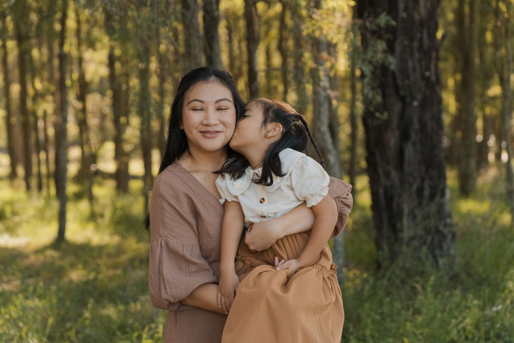 A mother and young daughter share a moment in a sunlit forest, the child whispering in her mom's ear. Family photoshoot in Rouse hill