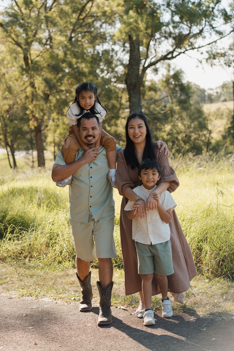 Happy family of four posing in a sunny park, father giving daughter piggyback ride with mother and son during Rouse hill family photoshoot
